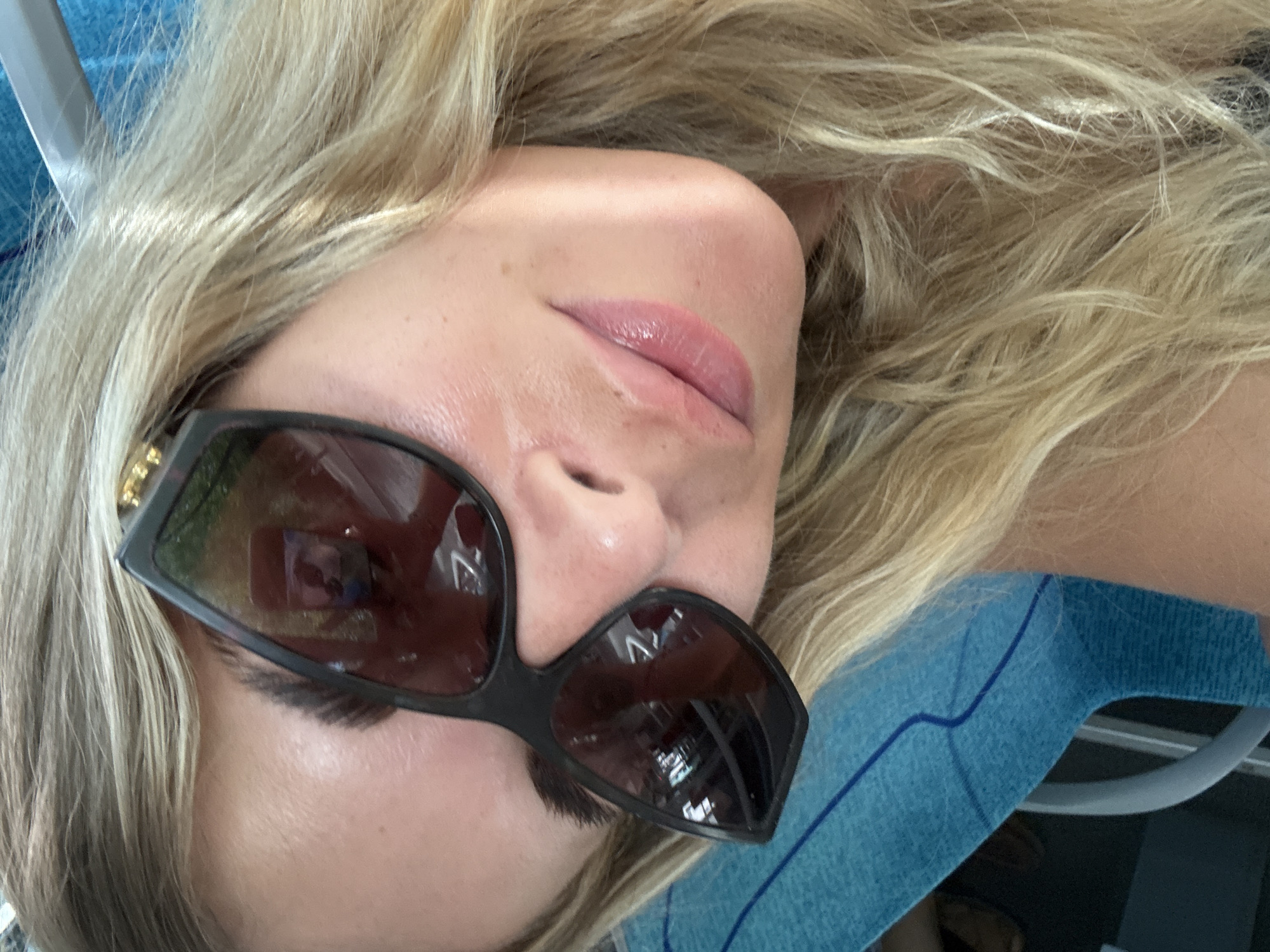 A close-up, angled, selfie-style shot shows a blonde woman wearing black, large-framed sunglasses. The woman's face is angled away from the camera. She has fair skin and is wearing pink lipstick. Her blonde hair is visible framing her face. The sunglasses reflect a scene, showing a blurred image of what appears to be a person holding a phone. The woman is wearing a blue garment with a dark blue line pattern, possibly a seat or clothing. The background is out of focus. The image was taken outdoors, possibly on a form of public transportation or in a seating area. There is no information or visual cues within the image to suggest it was taken in Paris, France.