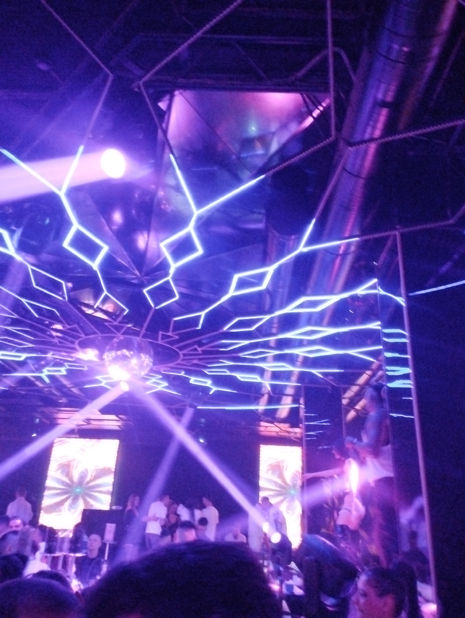 The image captures the vibrant interior of a nightclub or bar, characterized by an intense interplay of purple and blue lighting. The scene is dimly lit, creating a dynamic and energetic atmosphere.
**Subjects and Objects:**
The dominant feature is the ceiling, adorned with an intricate pattern of bright, glowing neon lights forming geometric shapes, primarily squares and rhombuses. A reflective disco ball is centrally placed on the ceiling, scattering light throughout the venue. Multiple spotlights emit powerful beams of white and purple light that cut through what appears to be a hazy or smoky atmosphere. Large, silver-toned ventilation ducts are visible on the ceiling, reflecting the colorful lights. Walls are lined with mirrored or highly reflective surfaces, which multiply the light patterns and reflections of the crowd, creating a complex visual effect. In the background, two large rectangular screens display abstract, swirling light patterns.
**People and Activity:**
A crowd of people is visible towards the lower and middle parts of the image. Their forms are largely obscured by the low light, motion blur, and reflections, but they appear to be standing, socializing, and enjoying the club environment. Some individuals are seen holding drinks, suggesting a social gathering or party.
**Notable Details:**
The predominant color scheme of purple, blue, and magenta bathes the entire space, giving it a modern and electrifying feel. The strong light beams and reflections contribute to a sense of depth and immersive visual experience. The setting is clearly indoors at night, relying entirely on artificial illumination. The overall mood is lively and engaging, indicative of a nightlife event. No discernible text is visible in the image.