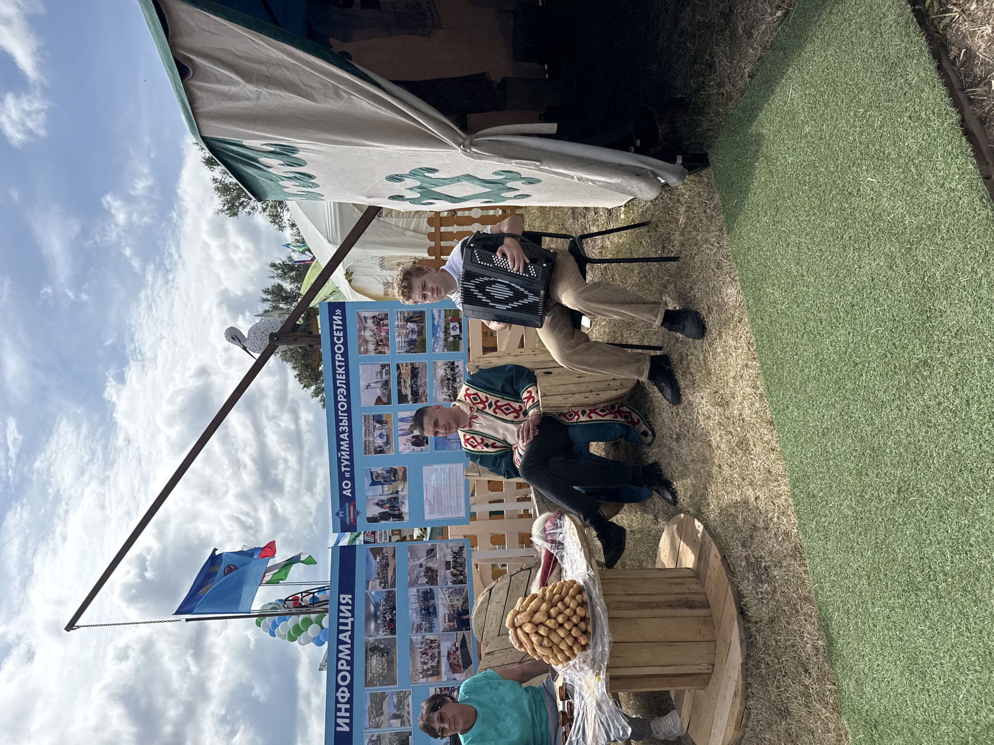 In the city of Nurkeevo, Russia, at an outdoor event, two men and a woman are gathered. A young man with curly blonde hair is seated on a black chair, playing an accordion. Beside him, a man in a traditional green and red embroidered robe is seated. To the left, a woman with glasses and a teal shirt is seated. In front of them is a wooden table made from spools, topped with a conical stack of pastries. Two informational boards are visible behind them, one displaying "ИНФОРМАЦИЯ" (Information) and the other advertising "АО «ТУЙМАЗЫГОРСЕТИ»" (Tuymazy Gorseti JSC). Balloons are clustered near a flag pole with several flags. A white bird sculpture is perched on a metal rod above them. A portion of a white tent with green trim is visible on the right. The ground is a mix of dry grass and a section of artificial green turf. The sky is cloudy with patches of blue.