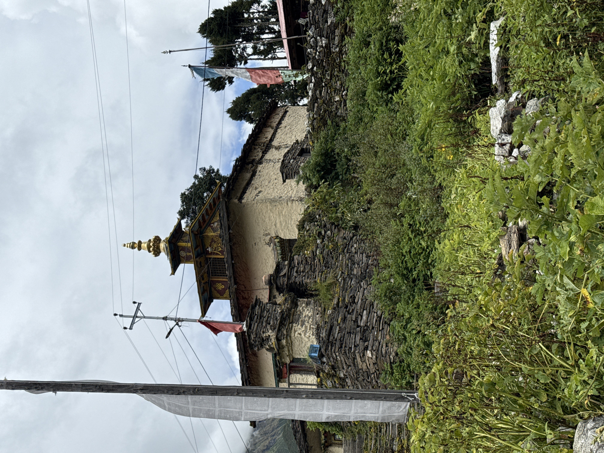 In Samagaon, Nepal, a traditional stone building with a golden pagoda-style roof stands on a hillside. The building's walls are made of rough, stacked stones, and it has a weathered, earthy appearance. A red flag flies from a stone pillar near the entrance, and a multi-colored flag, possibly representing a local or religious affiliation, waves from another pole. The structure is surrounded by dense green vegetation, including bushes, shrubs, and trees, with a cloudy sky overhead. Stone steps lead up to the building, indicating its elevated position. The overall scene conveys a sense of ancient architecture and natural beauty in a rural, mountainous setting.