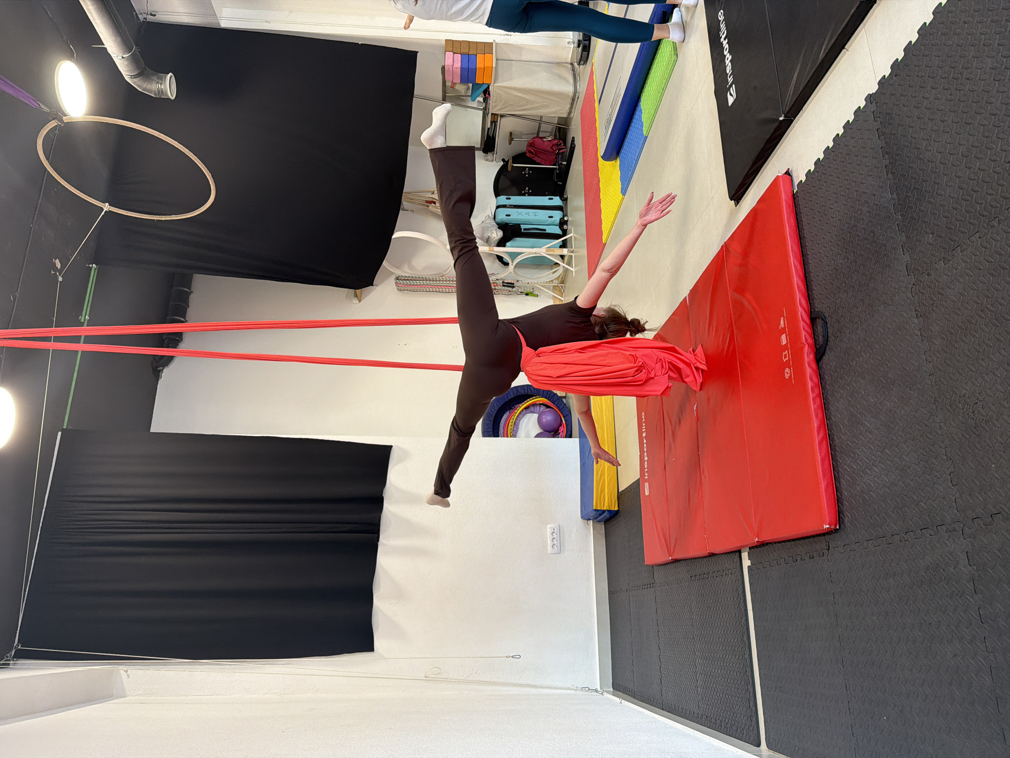 The image depicts an indoor aerial fitness studio in Bar, Montenegro, during an active training session. A woman, dressed in a dark brown one-piece outfit, is suspended upside down by red aerial silks, performing an acrobatic pose resembling a straddle or split. Her left leg extends upwards, while her right leg is bent, and her arms are outstretched.
The studio environment features various specialized equipment. A wooden aerial hoop (lyra) hangs to the left, while large red and black crash mats cover the floor directly beneath the aerialist, supplemented by black interlocking gym tiles. In the background, storage shelves are visible, holding items such as blue and yellow gym blocks, colorful hula hoops, and two light blue suitcases. A black fabric backdrop is draped along one wall. Bright artificial lighting illuminates the space from overhead fixtures. Another person's legs are partially visible in the upper right background. Text "InSPORTline" is visible on a black crash mat, and "XAH" appears on a blue suitcase.