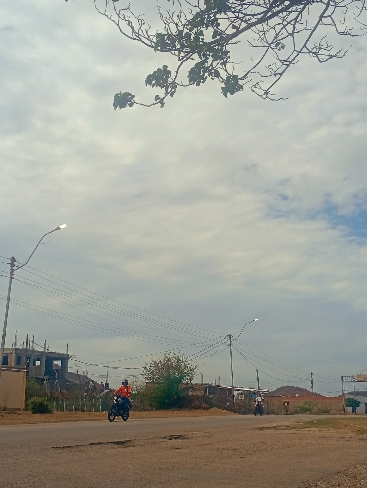 A full, low-angle outdoor shot captures a road in Altagracia, Venezuela, under a bright, cloudy sky. The foreground is dominated by a gray asphalt road with some patches and cracks. In the mid-ground, a motorcyclist wearing an orange shirt and a helmet rides a black motorcycle from left to right. Further back, another motorcyclist in a white shirt and black pants is also riding a motorcycle from left to right. The background features a partially constructed building on the left, with exposed rebar and concrete. There are utility poles with power lines spanning across the scene. Trees and smaller structures are visible further into the distance, with a low, hilly landscape in the far background. A tree branch with sparse green leaves hangs from the top of the frame.