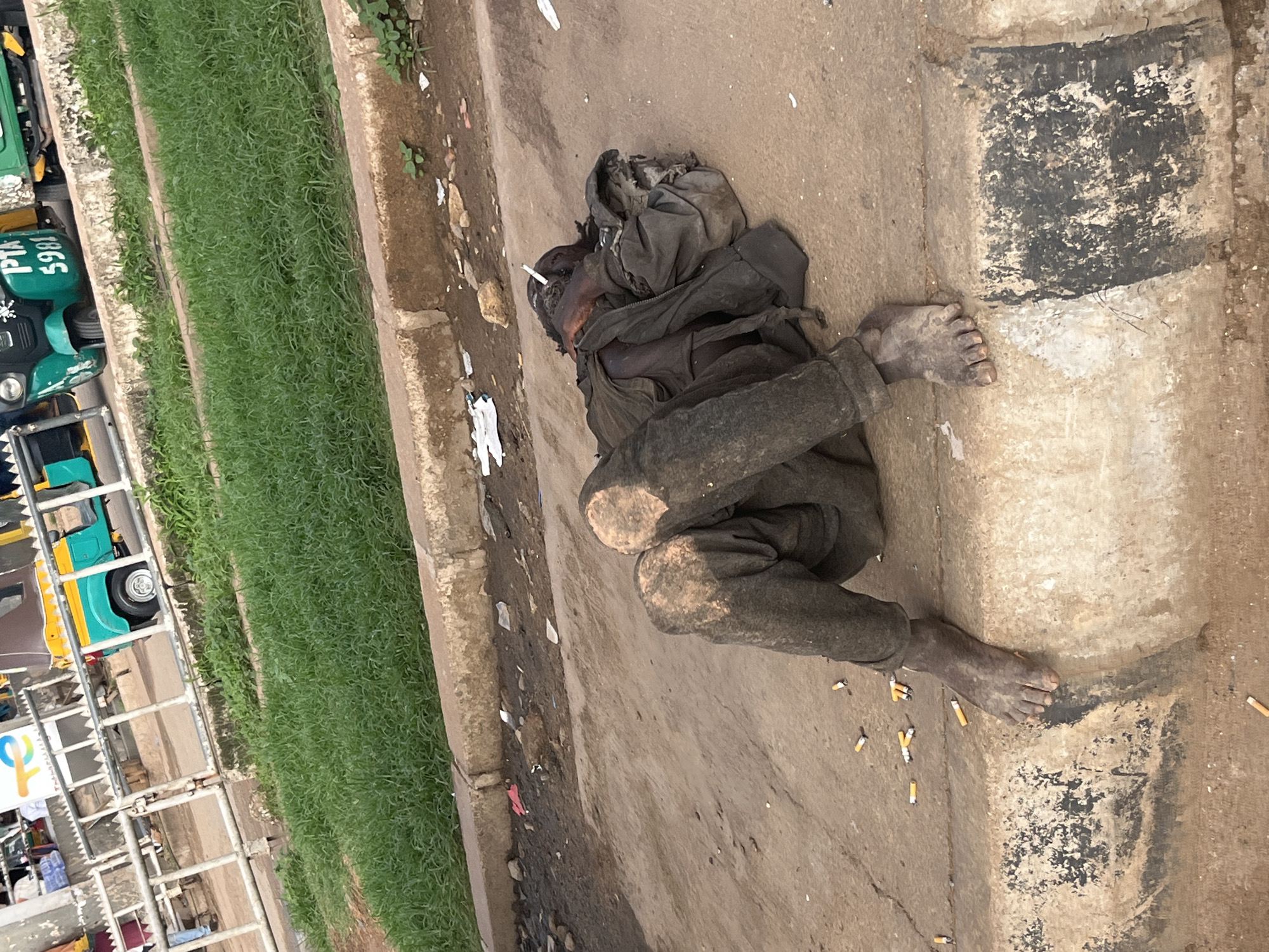 A person, appearing to be homeless, lies on a paved surface next to a curb in Gura Top, Nigeria. The person is covered in dirt and wearing dark, soiled clothing. Their legs are bent, and one foot rests on the curb. Several discarded cigarette butts are scattered around the person and on the curb. In the background, a green and yellow three-wheeled vehicle, possibly a tuk-tuk, is visible, along with some grassy areas and a metal railing. The scene appears to be a roadside or public area.