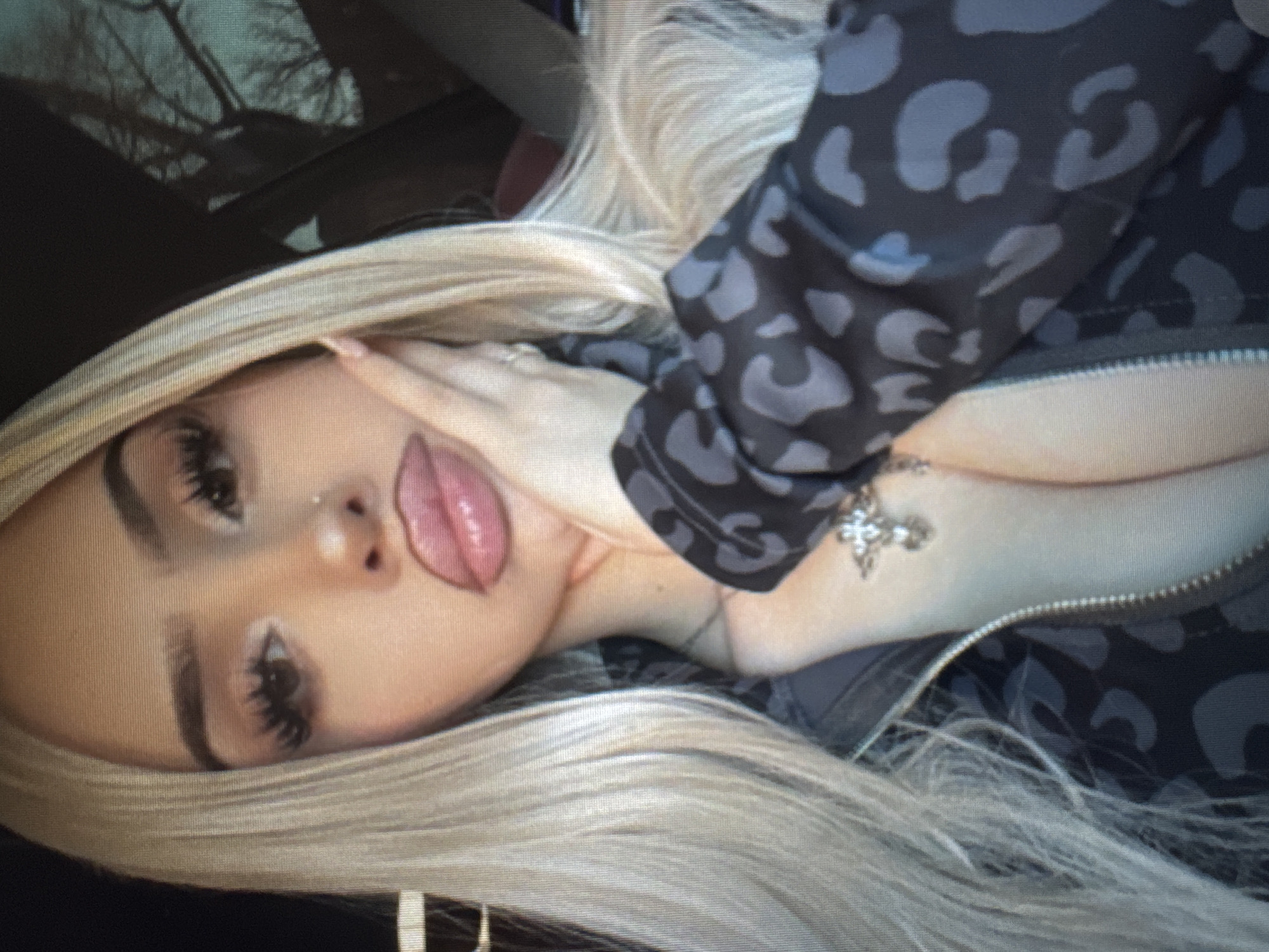 A close-up portrait captures a person, likely female, with long, platinum blonde hair. The person's head is tilted, and they are looking towards the viewer with a posed expression. They wear a dark grey zippered jacket featuring a repeating pattern of abstract, lighter grey leopard spots. A silver cross pendant is visible on a necklace around their neck.
The person's face shows prominent makeup, including dark, voluminous false eyelashes, sculpted eyebrows, and full, glossy lips that are slightly parted, revealing a portion of their tongue. A small, clear nose piercing is also visible. One hand is gently placed with fingers resting near their cheek and jawline.
The background is dark and blurred, suggesting an out-of-focus environment. It appears to show indistinct shapes that could be bare tree branches or reflections against a dark surface, possibly seen through a window, such as from inside a vehicle or a dimly lit room. The lighting is soft but does not clearly indicate a specific time of day. No discernible text is present in the image.