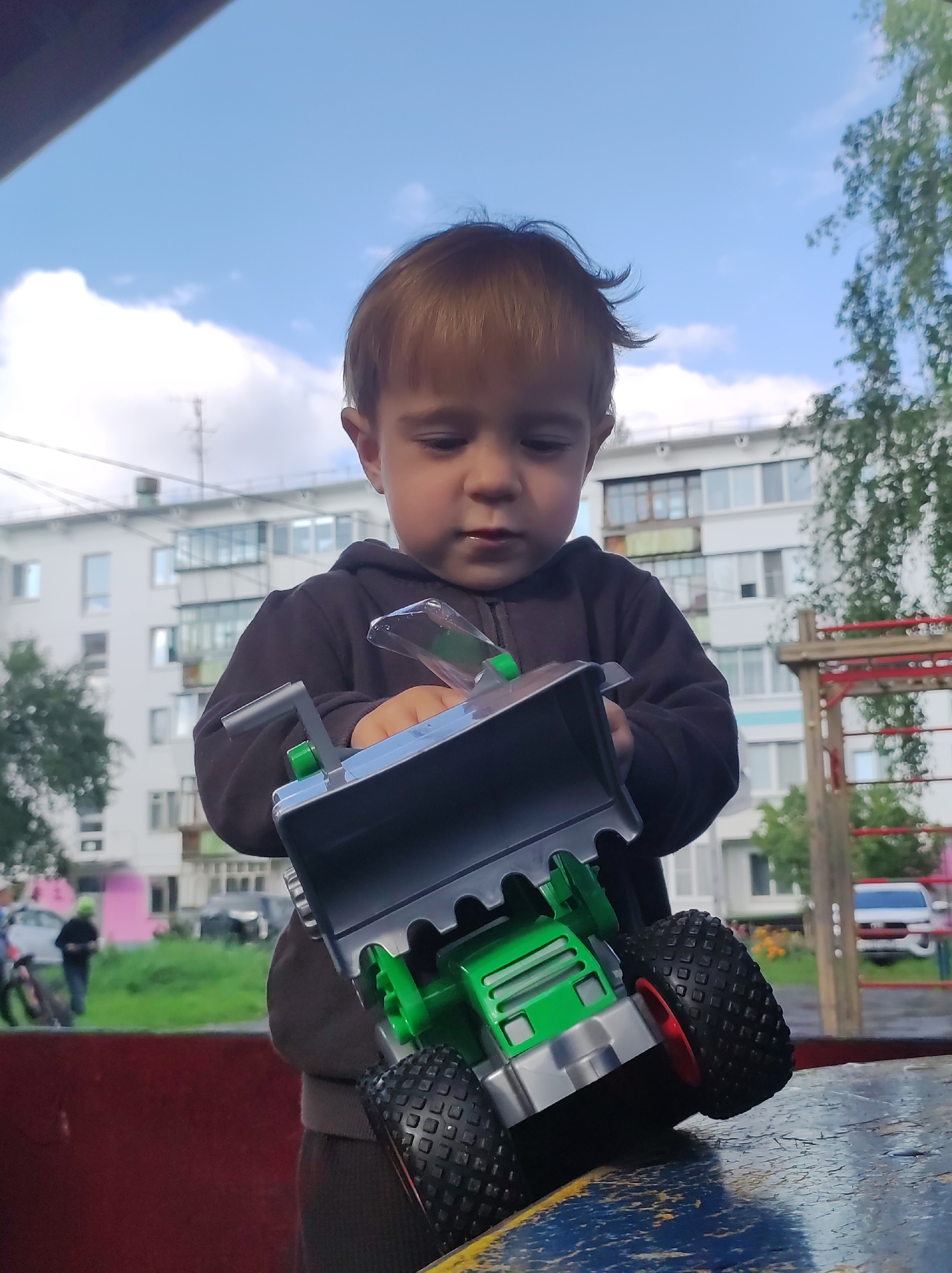 An outdoor, low-angle, medium shot captures a young Caucasian child playing with a toy truck. The child, with light brown hair, is wearing a dark hooded sweatshirt and is focused on manipulating the toy, which is primarily green and gray with large black tires and red rims. The child is standing on what appears to be a wooden surface, possibly part of a playground.
In the background, a residential building with multiple floors and balconies is visible. Trees with green foliage are present, suggesting a park or residential area. A playground structure, red and wooden, is also in the background. The sky is blue with scattered white clouds. The scene appears to be in a city in Russia, specifically Chaikovsky, Russia, indicated by the typical Soviet-era apartment block architecture. The overall atmosphere is that of a casual, everyday moment of childhood play.