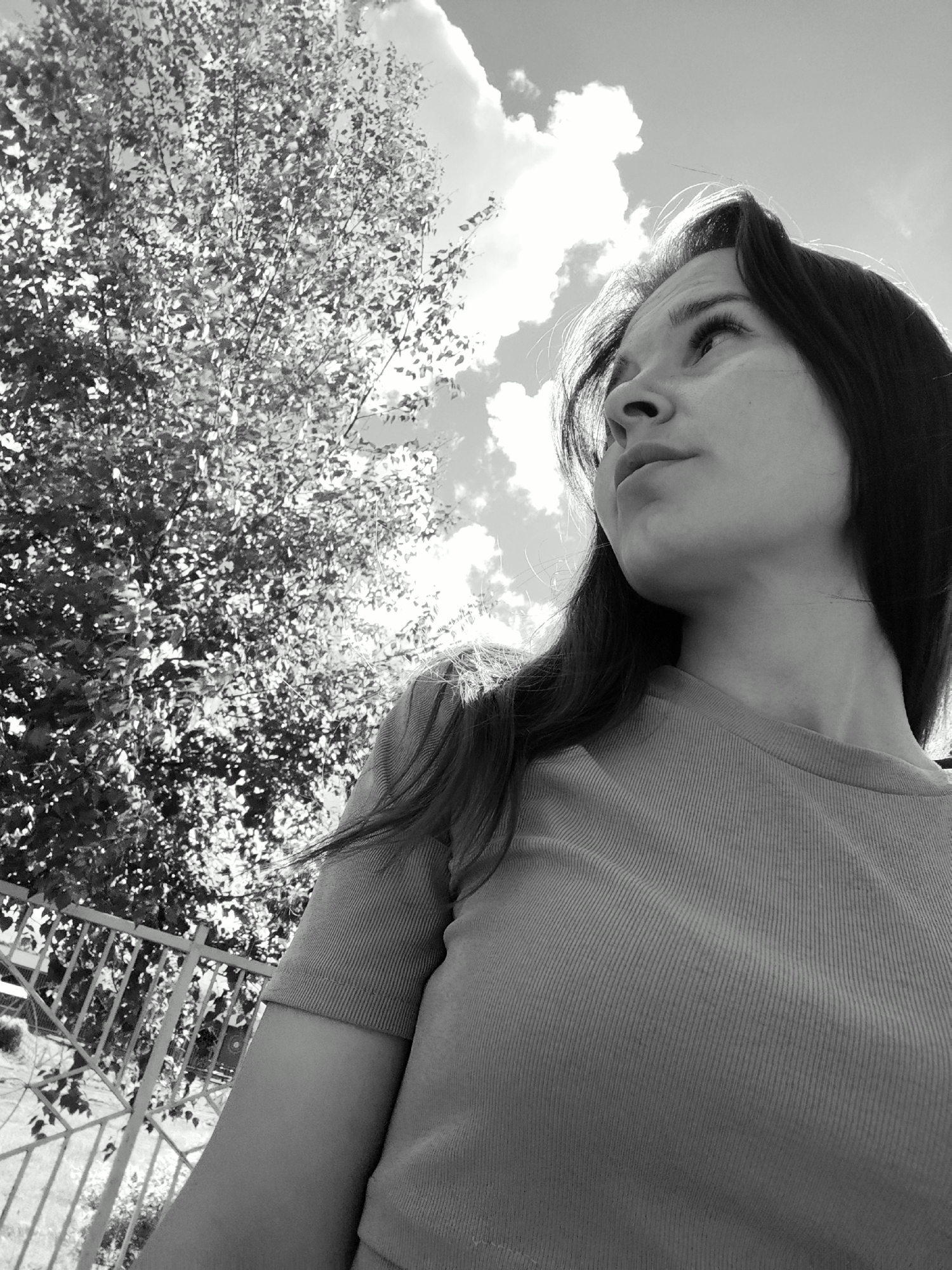This black and white photograph captures a low-angle perspective of a young woman looking upwards and to her right, against a brightly lit sky filled with fluffy clouds. Her profile is prominent in the foreground, with her long, dark hair falling over her shoulders and back. She wears a simple, short-sleeved top with a subtle ribbed texture. Her expression appears calm and observant, her gaze directed towards something unseen above.
To the left and behind her, a large tree with dense foliage occupies much of the background. The sunlight filters through its leaves, creating a high-contrast effect with numerous bright highlights, characteristic of a sunny day. A metal fence with horizontal and vertical bars is partially visible in the lower-left corner, suggesting an outdoor setting like a garden or park in Fokino, Russia. The sky is largely clear, save for the distinct, bright clouds. The black and white conversion emphasizes the textures of the leaves, clouds, and the woman's clothing and hair, as well as the strong interplay of light and shadow, indicating a bright midday or afternoon sun. There is no visible text in the image.