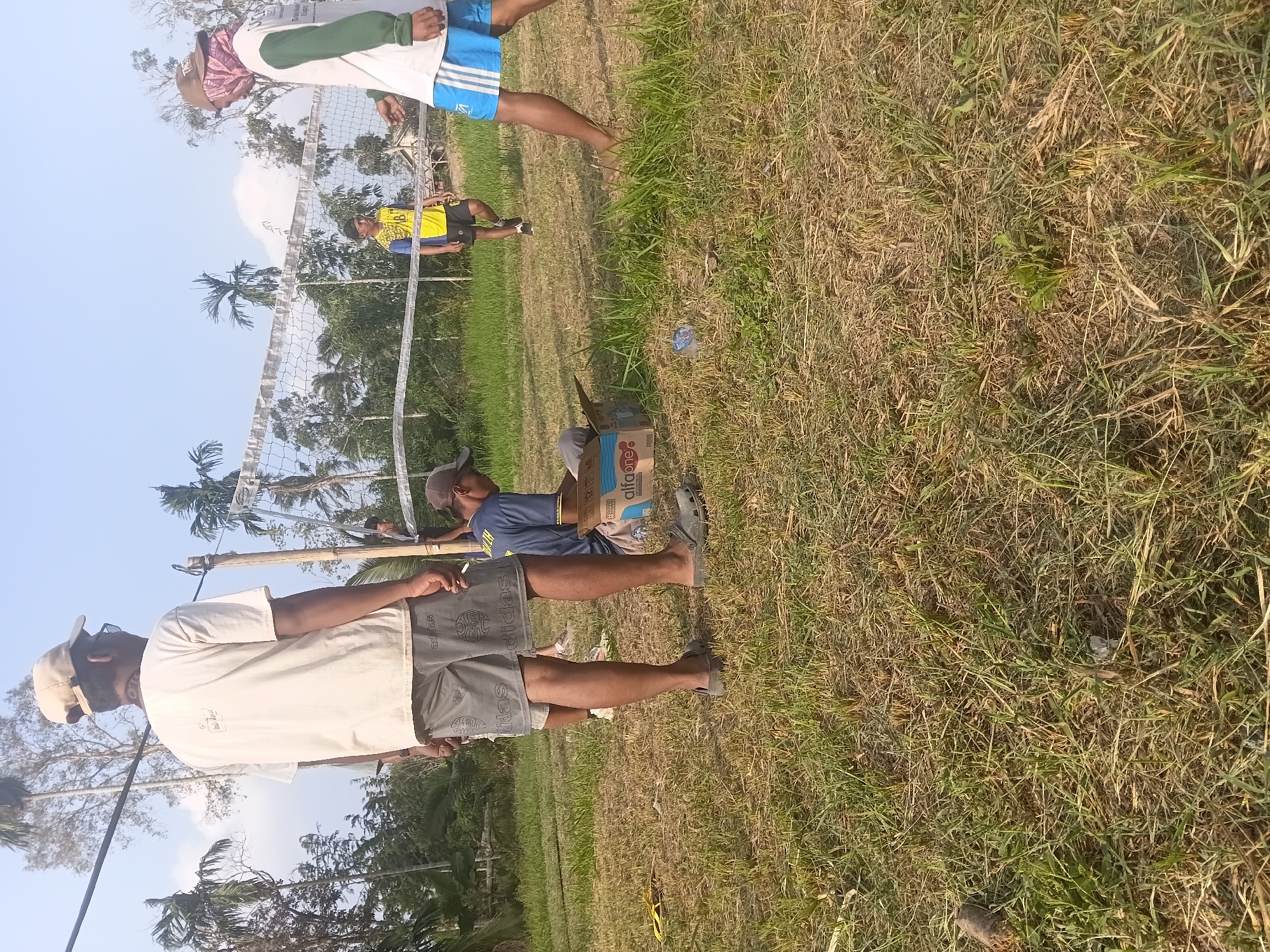 The image depicts an outdoor scene in Margo Rukun, Indonesia, characterized by a grassy field and a volleyball net. In the foreground, a person wearing a beige cap, a white t-shirt, and grey shorts stands facing away from the camera. To their left, a person in a blue shirt and a brown cap is seated on the ground, looking into an open cardboard box. Further in the background, another individual in a yellow and blue shirt is positioned near the volleyball net, and a person in a white t-shirt with green sleeves and blue shorts is on the right side of the frame, facing the net. Trees and vegetation surround the open field, under a bright, clear sky. The ground is covered in dry grass, with some patches of greener growth. A plastic bottle and a yellow flip-flop are visible on the ground near the foreground.