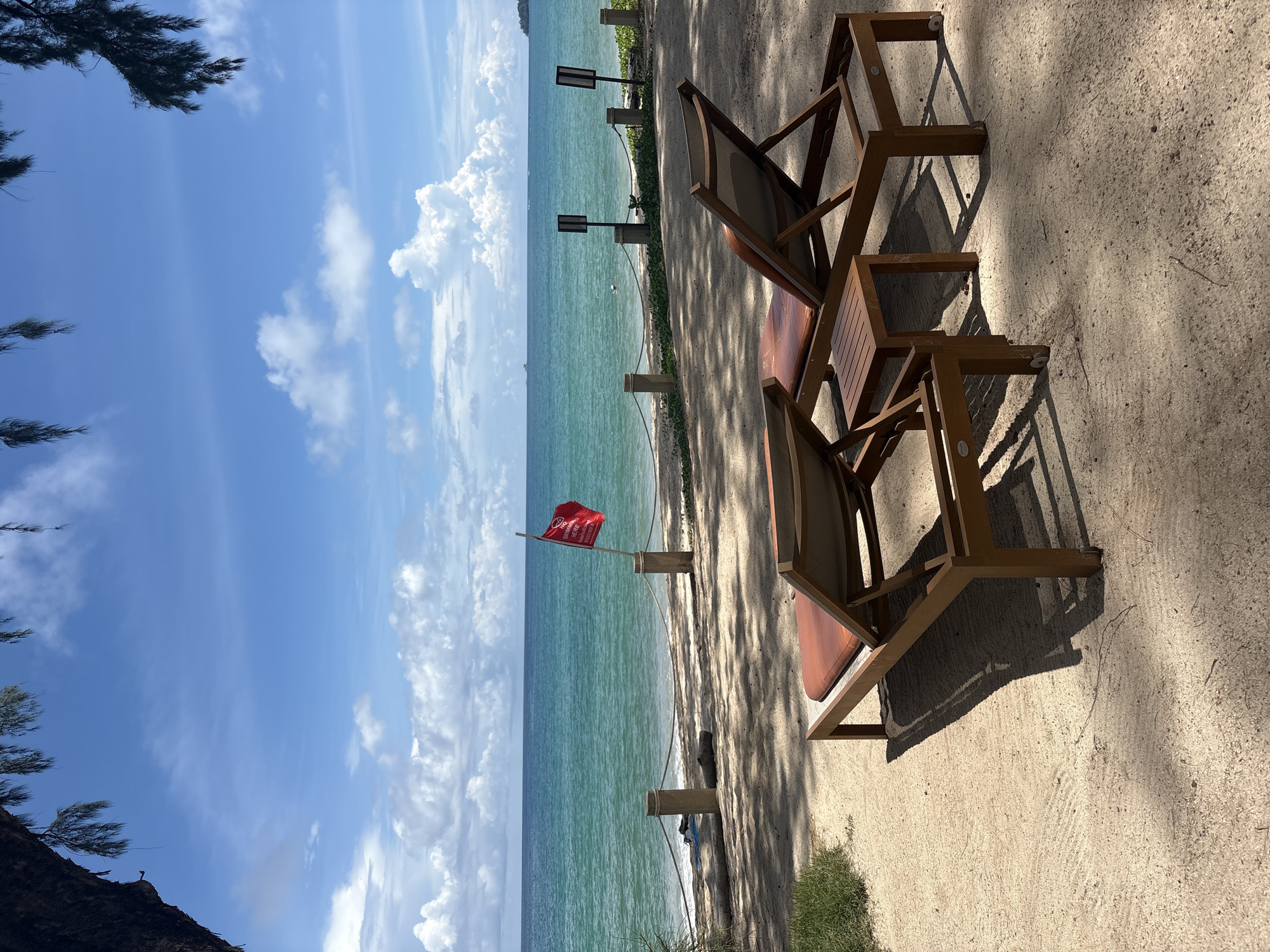 Two wooden lounge chairs with orange cushions are positioned on a sandy beach in Choeng Thale, Thailand. A small wooden side table sits between the chairs. In the background, the turquoise ocean meets a bright blue sky with scattered white clouds. A red flag with white text is planted in the sand near the water, indicating a no-swimming zone. A rope barrier is strung between wooden posts along the edge of the beach. Palm fronds hang from the upper left corner of the image. The scene is shaded by trees, casting dappled shadows on the sand.