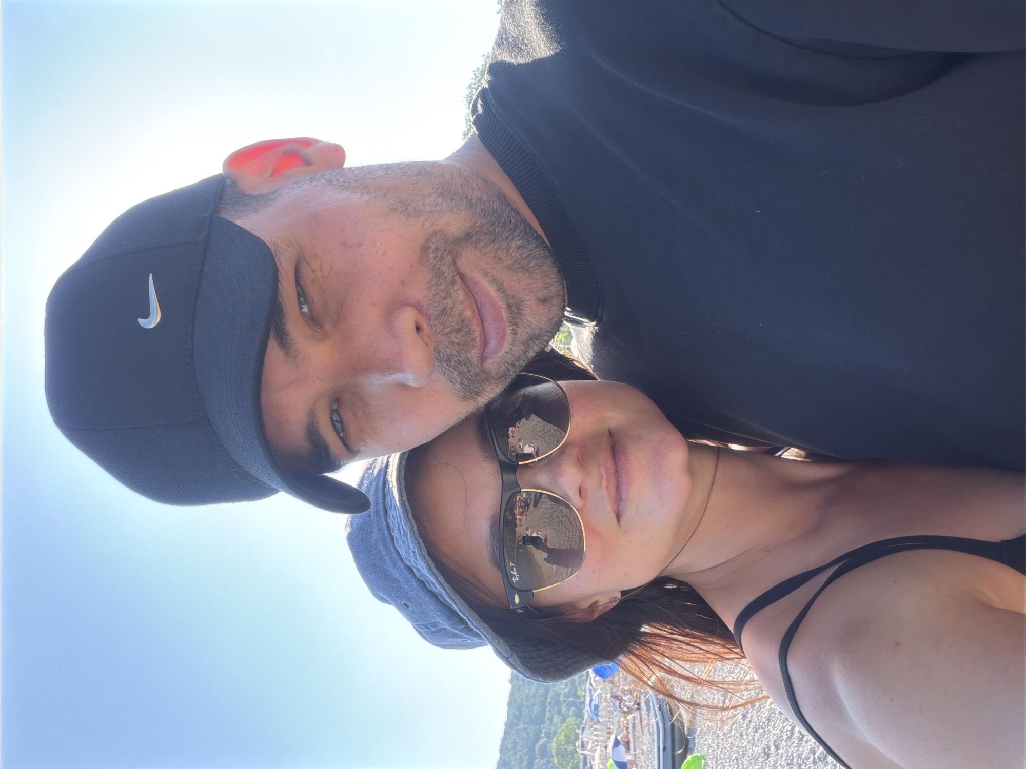 A couple poses for a selfie on a beach. The man, wearing a black Nike baseball cap and a black sweatshirt, is on the right. The woman, wearing a blue bucket hat and sunglasses, is on the left. The background shows a beach with pebbles, umbrellas, and mountains in the distance. The image was taken in Prijevor, Montenegro.