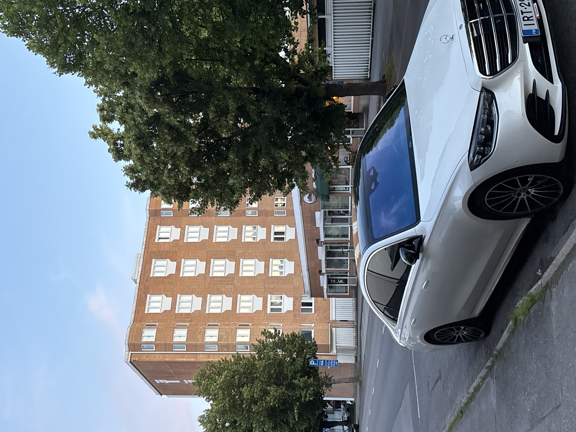 In the city of Pori, Finland, on a street with a paved road and sidewalk, a white Mercedes-Benz sedan is parked. The car is facing forward, its front quarter visible. Behind the car, a multi-story brick building with numerous windows stands. Trees with green foliage are positioned in front of the building and to its left. A street sign with a parking symbol is also visible near the building and trees. The sky above is clear with a light blue hue.