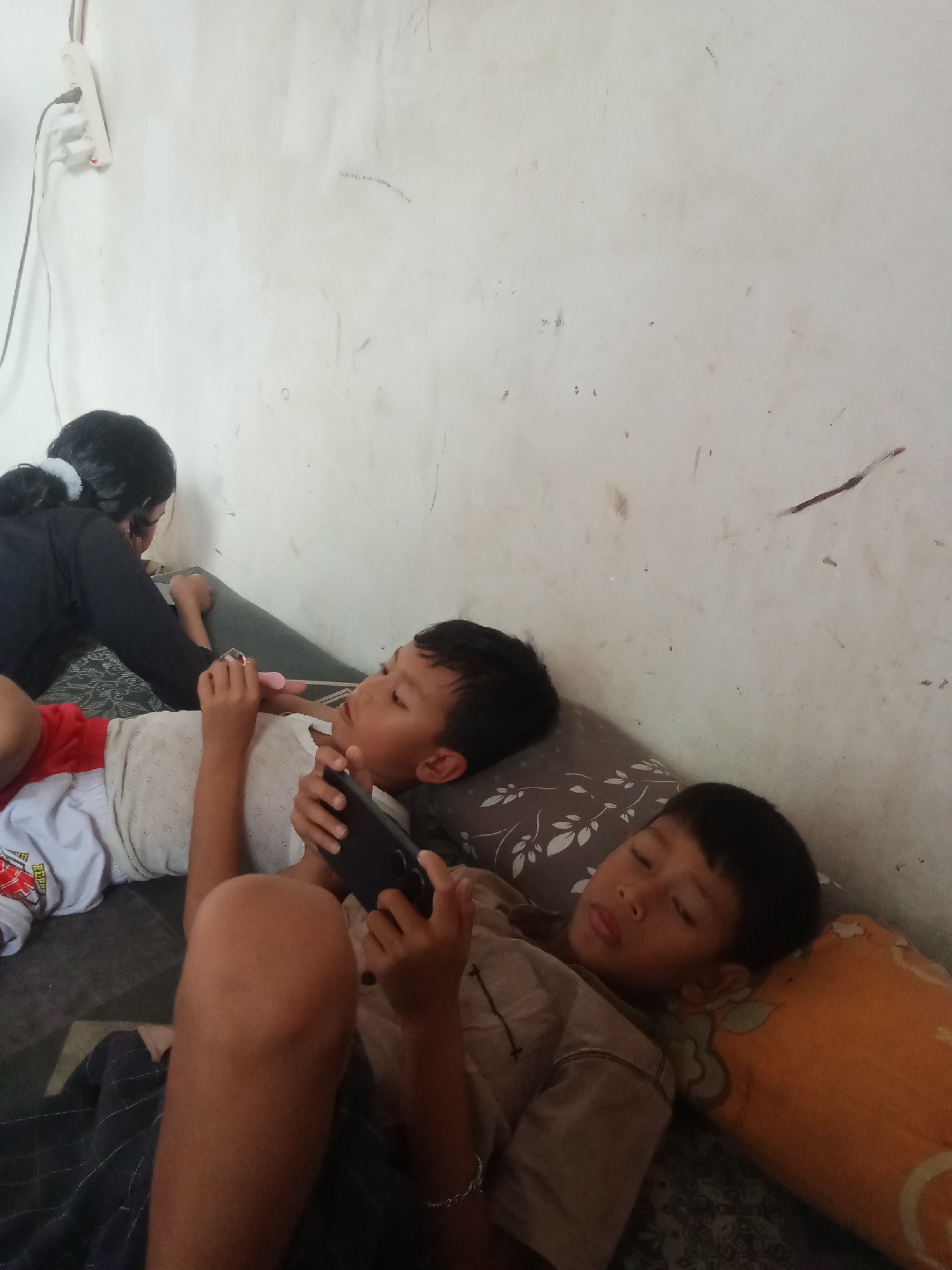 In what appears to be a residential interior in the city of Mojokerto, Indonesia, three individuals are depicted in a relaxed setting, likely a bed or sofa. On the left, a person with dark hair pulled back in a ponytail, wearing a black top, is leaning over, possibly interacting with a mobile device. In the center and right, two young boys are lying down. The boy in the center, wearing a white shirt with red accents, is looking towards a mobile device held in his hands. The boy on the right, wearing a brown shirt, is lying on his side, eyes closed, appearing to be asleep, with his head resting on an orange pillow. The environment is characterized by a light-colored, textured wall and what seems to be a patterned mattress or bed covering. An electrical outlet with a cord is visible on the upper left portion of the wall. The lighting suggests an indoor environment with ambient light.