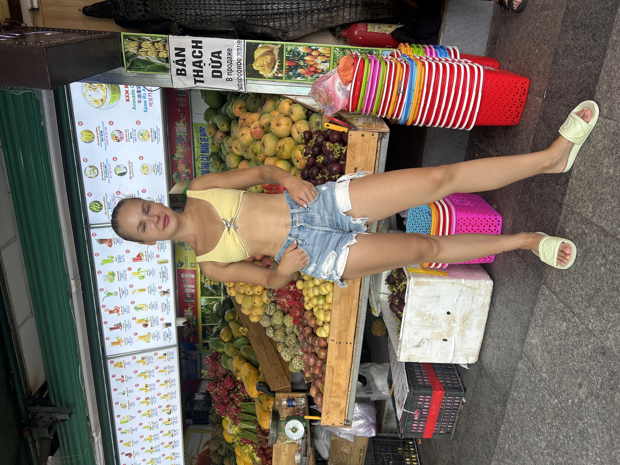 A young Caucasian woman stands in front of a fruit stand in Nha Trang, Vietnam. She is wearing a yellow bikini top and ripped denim shorts. The fruit stand displays a variety of fruits, including mangoes, dragon fruit, and mangosteen. The background shows a menu board with pictures of juices and ice cream. To the right of the woman, there are stacks of colorful plastic baskets and a large faux crocodile. The ground is paved with gray tiles.
