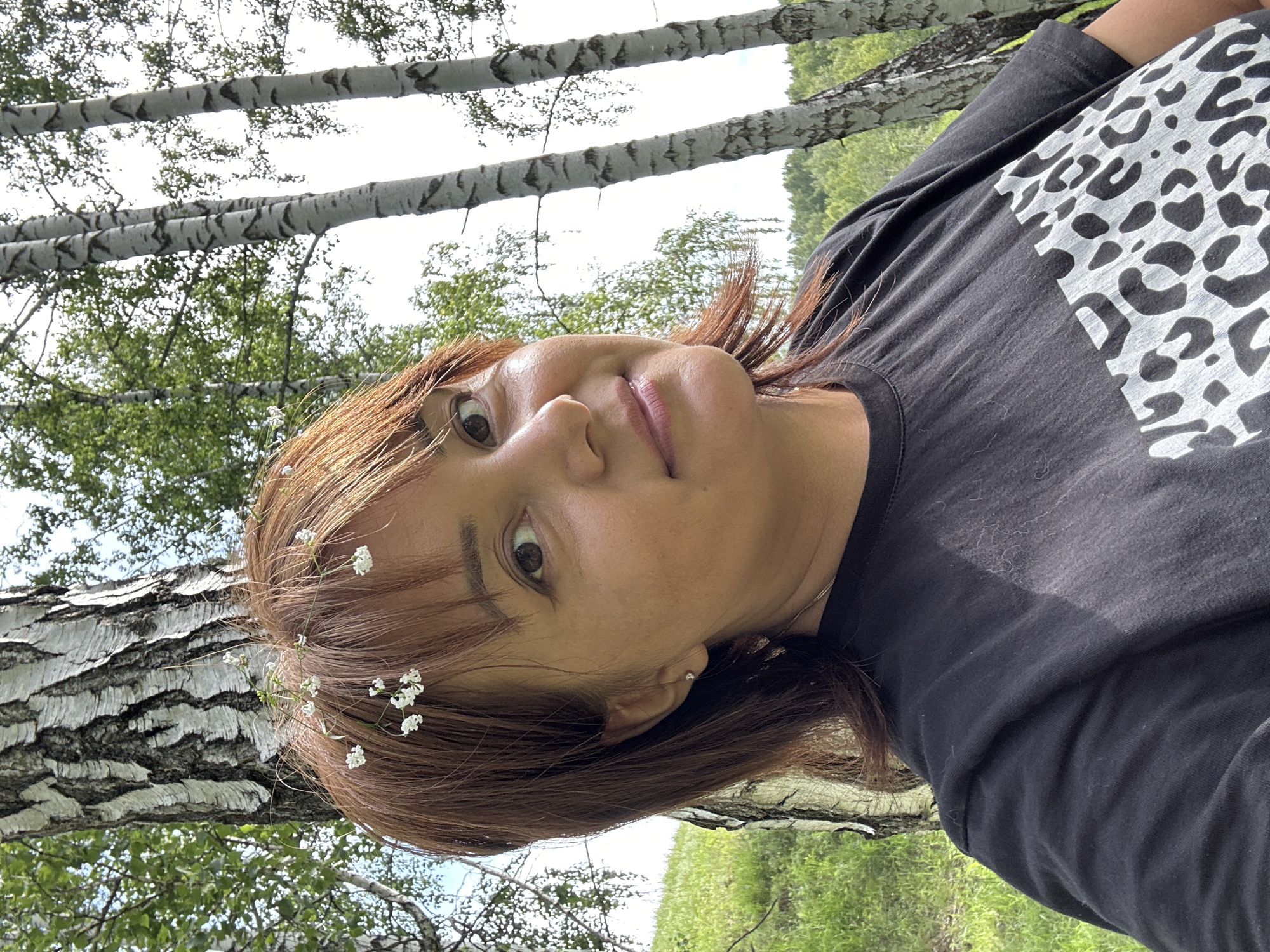 The image is a portrait of a woman in an outdoor setting, with birch trees and greenery in the background. The woman is looking up at the camera, with her head tilted. She has brown hair, which is adorned with small white flowers. She is wearing a dark t-shirt with a white leopard print design on the upper chest. Her expression appears to be relaxed and perhaps a little curious. The lighting suggests it is daytime, likely with natural sunlight filtering through the trees. The overall mood is serene and natural. There is no visible text in the image.