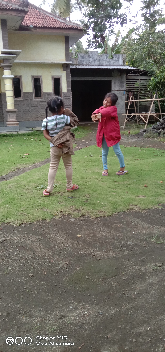 The scene depicts two young girls in an outdoor residential setting in Ranugedang, Indonesia. The foreground consists of a dark, gravelly dirt path. Moving backward, a rectangular patch of manicured green grass serves as the immediate ground for the subjects.
On the left, a girl with dark hair styled in two pigtails stands facing away from the camera. She wears a horizontal striped t-shirt (white and light green) and light tan leggings. Pink and green flip-flops are visible on her feet. She is holding a brown, patterned garment, draped over her shoulders and held behind her back.
To her right, a second girl, facing slightly left towards the first girl, has short dark hair and a smiling expression. She is wearing a long-sleeved red jacket and light blue jeans. She has a purple flip-flop on her left foot and a darker, patterned flip-flop on her right foot. Her arms are bent at the elbows, extended forward with open hands and splayed fingers, suggesting a playful pose.
The background features a light-yellow house with a reddish-brown tiled roof on the left, exhibiting decorative pillars and three rectangular dark-framed windows. Adjacent to this house on the right is a simpler structure, possibly unfinished, with a dark open doorway and a corrugated metal roof. Beyond these structures and above them, dense green vegetation, including palm trees and other foliage, is visible under a bright, overcast sky. To the far right, a wooden, scaffold-like structure stands near a pile of dark debris, and a small, light-colored bird is discernible on the ground.
