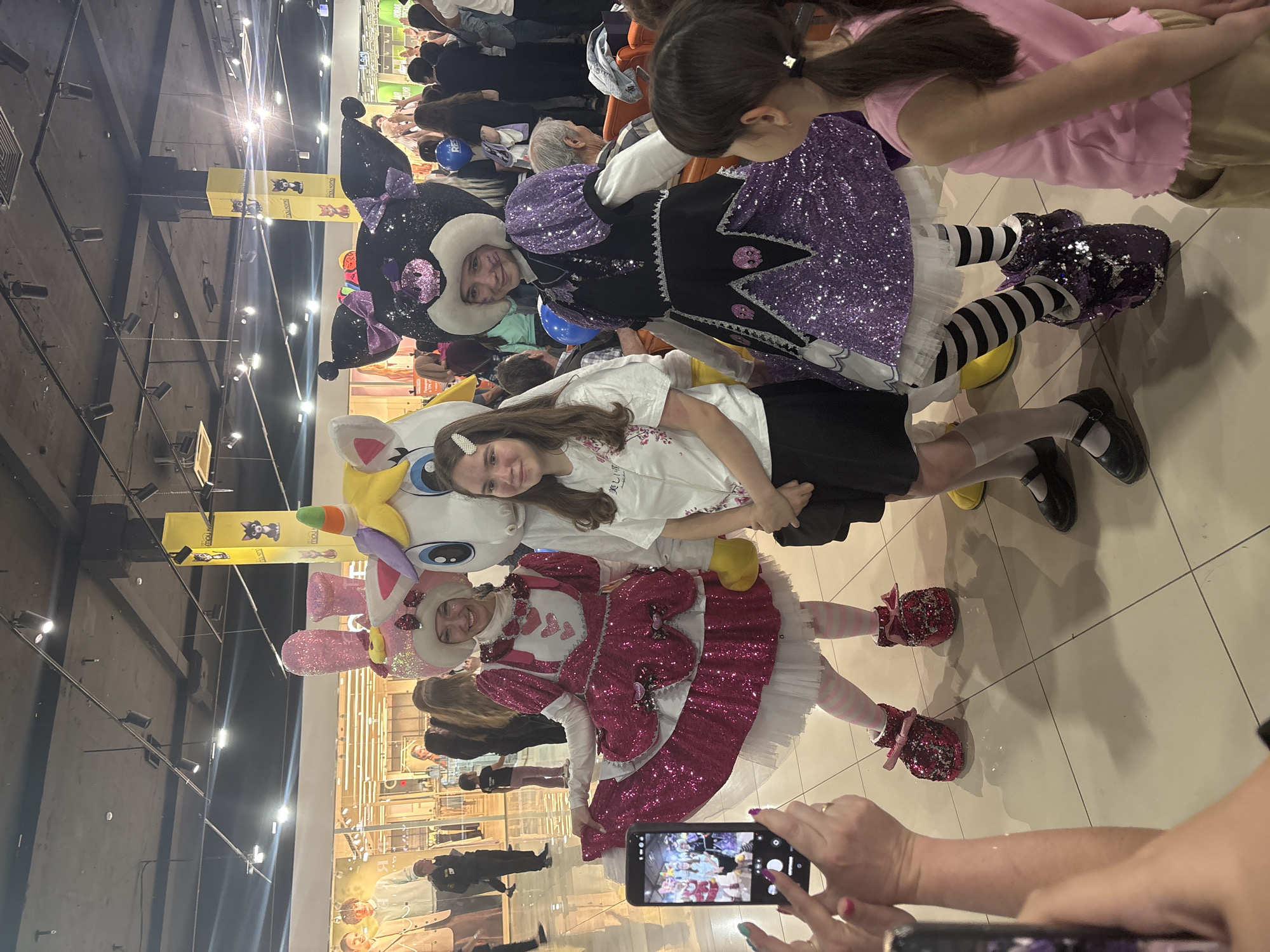 A medium shot captures a group of people posing for a photo in what appears to be an indoor public space, possibly a shopping mall in Tri Protoka, Russia. A young girl in a white t-shirt and black skirt is centrally positioned, leaning against a plush unicorn toy. To her left, a person is dressed in a pink, sequined outfit with bunny ears, and to her right, another person is wearing a black and purple sequined costume with a large collar. Both costumed individuals are smiling.
In the lower foreground, a person's hands hold a smartphone, seemingly taking a photo of the group. In the background, other people are visible, suggesting a crowd. The mall's interior features a polished floor, overhead lighting, and large advertisement boards on the wall.
