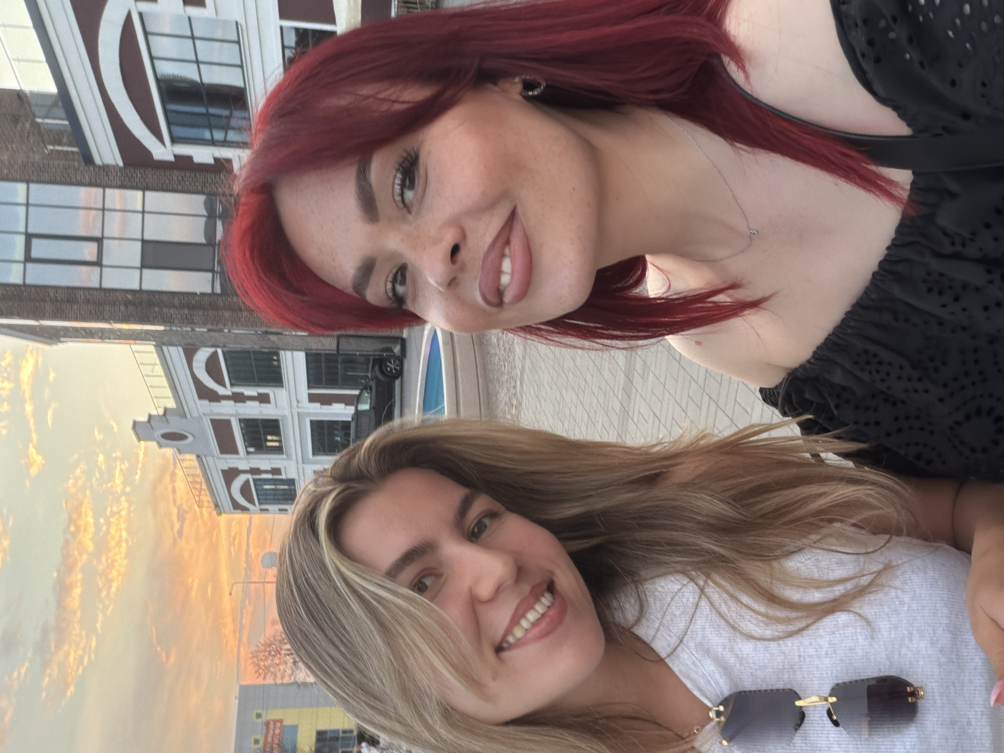 Two women are taking a selfie outdoors in the city of Tolyatti, Russia, with buildings and a sunset visible in the background. The woman on the left has blonde hair and is wearing a white top and sunglasses. The woman on the right has vibrant red hair and is wearing a black off-the-shoulder top. They are both smiling at the camera. The environment appears to be a public space with paved ground and modern architecture. The sky is filled with warm-toned clouds, suggesting it is either sunrise or sunset.