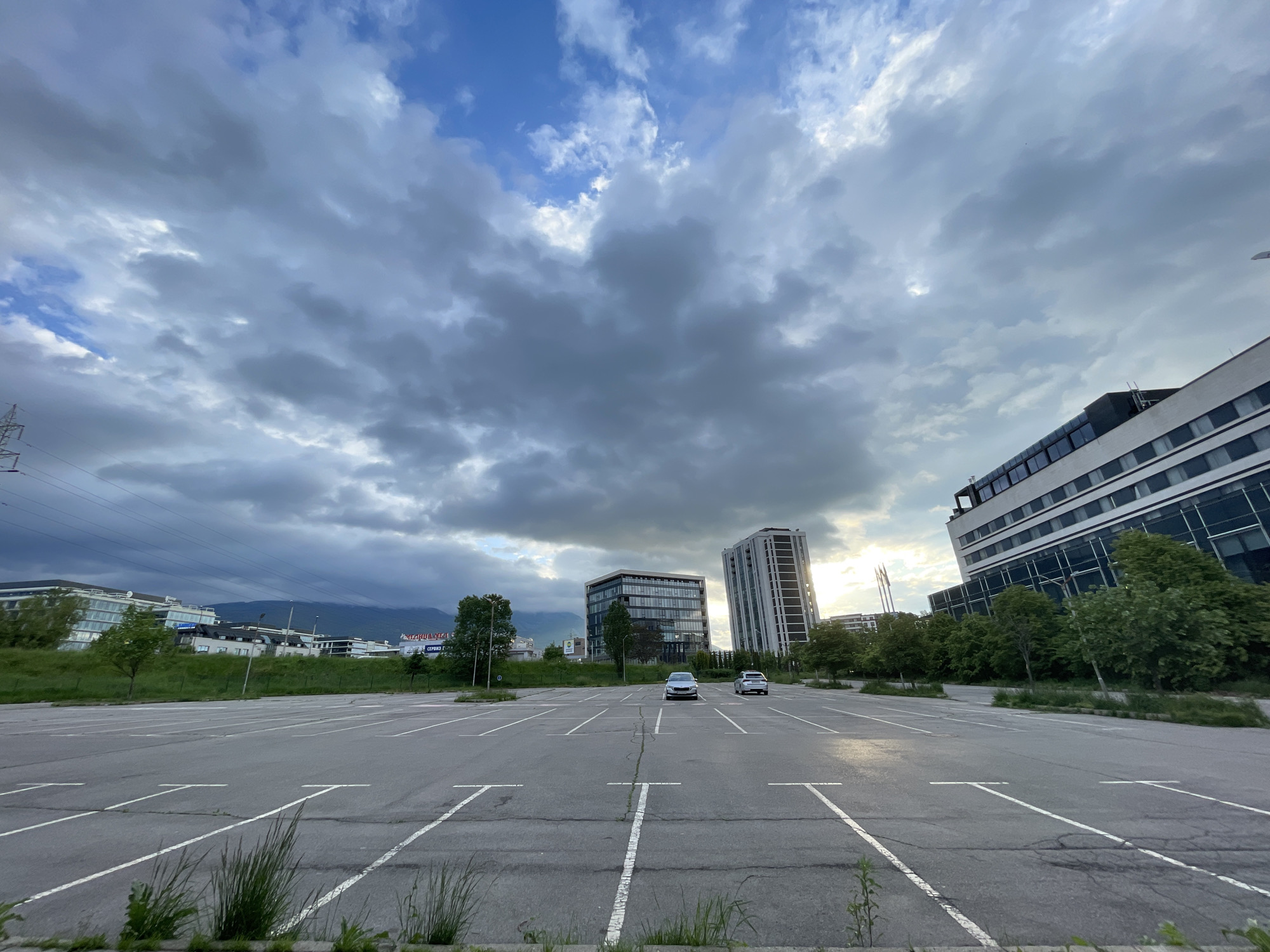 The image depicts an expansive, mostly vacant parking lot in Sofia, Bulgaria. Two light-colored cars are visible, parked in the middle of the lot. The parking spaces are marked with white lines, many of which are faded and cracked. Vegetation, including grass and small weeds, grows sparsely in the cracks and along the edges of the parking area. In the background, modern office buildings and a tall, possibly residential, building form a skyline. Trees are interspersed among the buildings. A mountain range is visible in the distance. The sky is dominated by heavy, overcast clouds, with patches of blue sky peeking through. A power transmission tower is visible on the left.