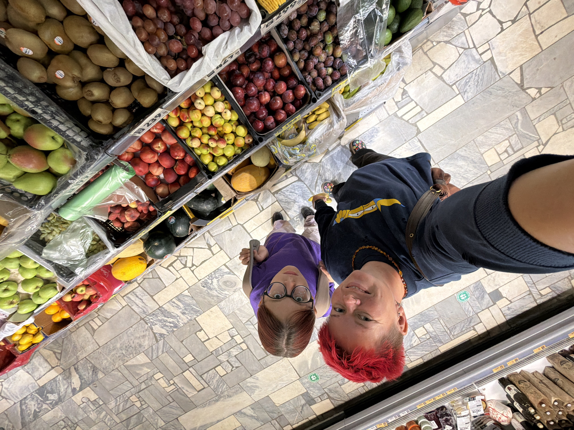 The image is a high-angle, full-body shot of two people in what appears to be a grocery store produce section. The person in the foreground, on the bottom right, has bright red hair and is wearing a dark blue sweatshirt with a yellow cartoon character on it. They are holding a phone in their right hand, taking the selfie. To their left and slightly behind is another person with dark red hair, wearing glasses and a purple t-shirt. They are looking up at the camera. Both individuals are standing on a patterned tile floor. In the background, rows of various fruits are visible in black plastic crates, including apples, pears, kiwis, grapes, plums, bananas, and what appears to be a melon. The scene is brightly lit, suggesting an indoor environment.