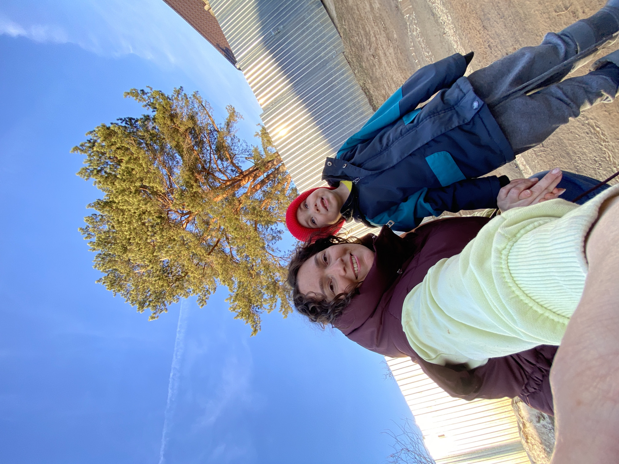 A selfie taken from a low angle captures a woman and a child in an outdoor setting in the city of Chaikovsky, Russia. The woman, with curly brown hair, wears a maroon puffer vest over a light green top. Next to her, a child with a red hat is bundled in a navy and teal jacket and gray pants. The child holds the woman's hand. Behind them, a tall pine tree with yellow-green foliage reaches up towards a clear blue sky with a contrail. In the background, there is a corrugated metal wall and a glimpse of a building's roof. The lighting suggests it is daytime.