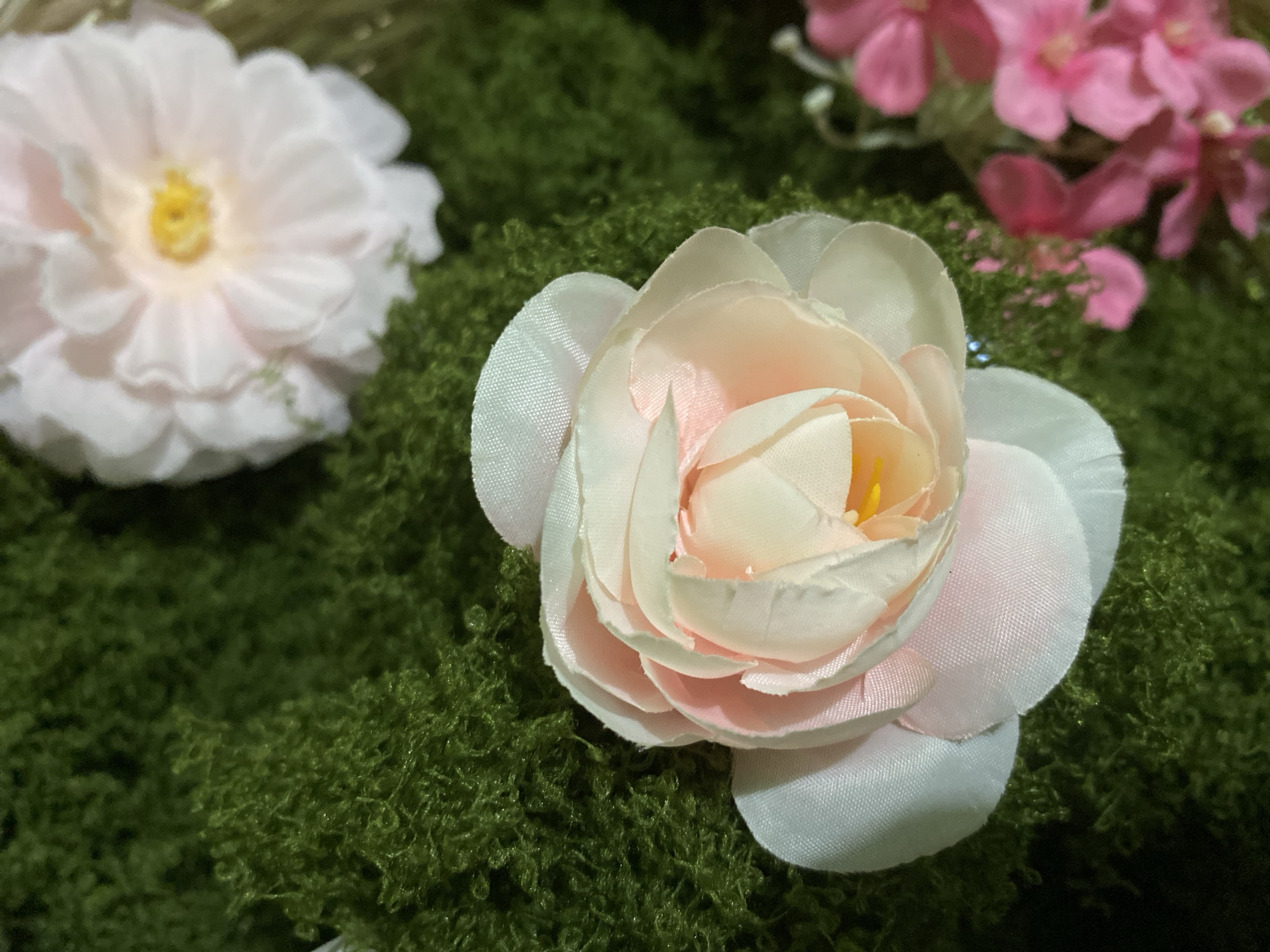 This media file depicts a close-up of artificial flowers arranged on a bed of green moss. The primary subjects are two light pink and white roses, with a background cluster of pink flowers. The rose in the foreground is partially unfurled, revealing layers of delicate petals in shades of cream and pale pink, with a bright yellow center. The rose on the left is fully bloomed, showcasing a similar petal structure and a soft yellow center, appearing slightly out of focus. In the upper right corner, several smaller pink flowers with darker centers are visible, also artificial. The entire arrangement is set against a textured green moss, which provides a naturalistic base for the decorative flowers. There is no visible text, and the lighting suggests an indoor setting, possibly for decoration or a craft project. The scene evokes a sense of artificial beauty and floral arrangement, with no specific activity or event taking place, nor any environmental cues like time of day or weather. The location context provided is Kuala Pembuang, Indonesia, but this information does not translate into visible cues within the image itself.
