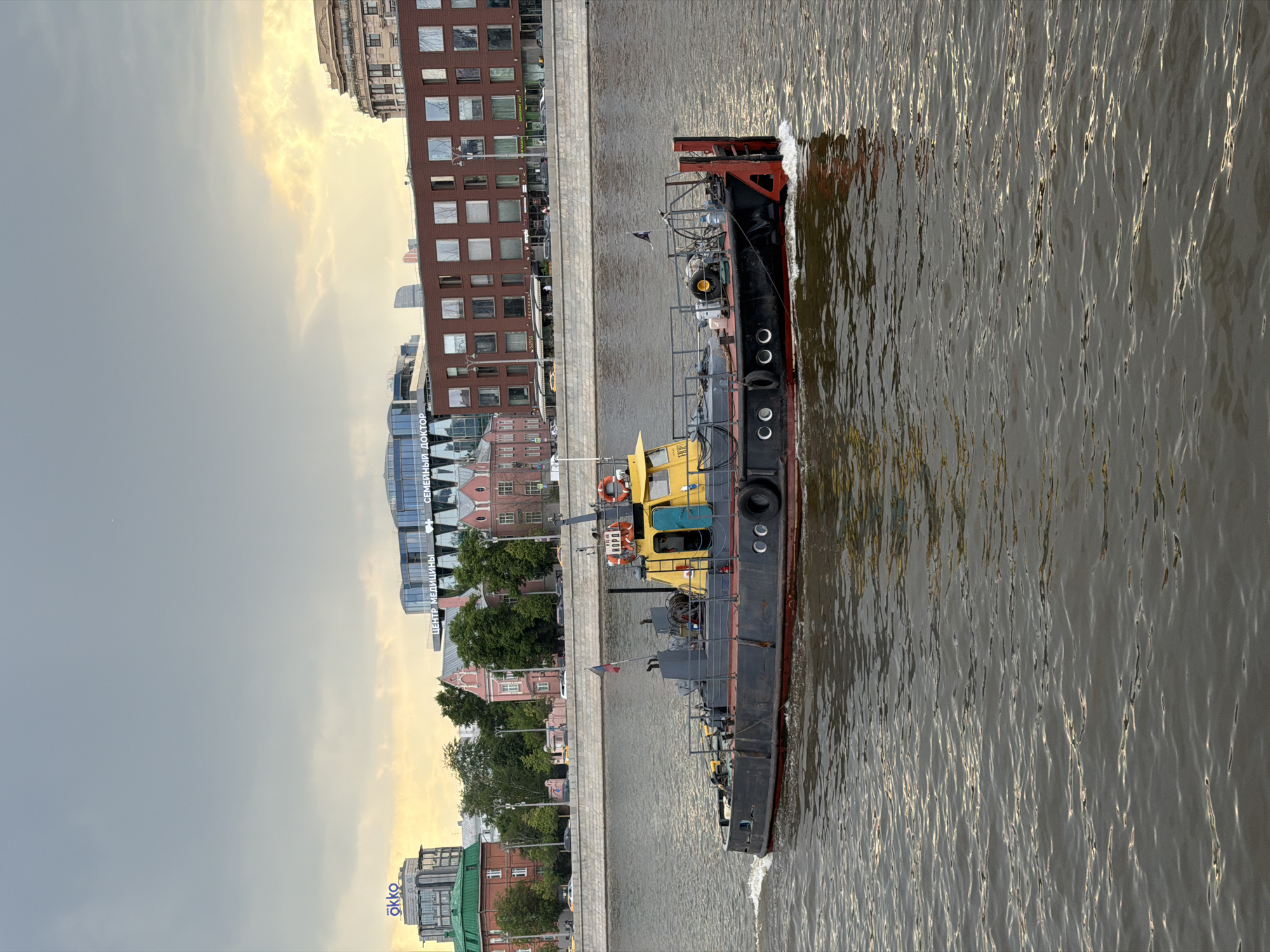 An eye-level, outdoor shot shows a yellow and black tugboat traveling in the Moskva River in Moscow, Russia, with buildings along the riverbank.
The tugboat, seen from its port side, has a yellow upper section and a black lower section with red highlights on the stern. It is traveling toward the upper left of the frame, creating ripples in the water.
The riverbank to the left of the boat is lined with a concrete embankment. Beyond the embankment are various buildings, including a large red brick building with multiple windows, modern glass-fronted structures, and older buildings with trees in front of them.
The sky is overcast with a mix of gray and yellow hues, indicating either early morning or late afternoon light.