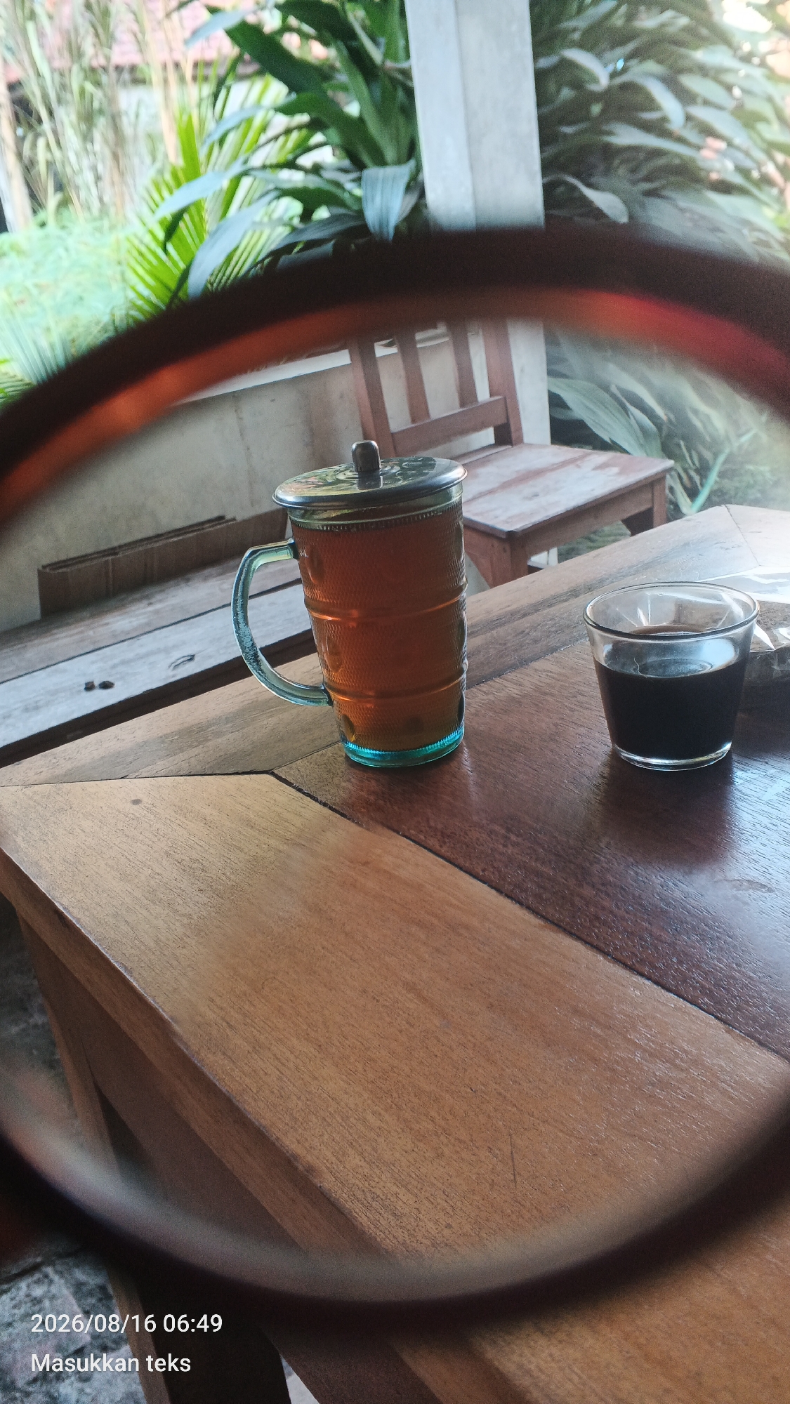 A low-angle, close-up shot captures two drinks on a wooden table, viewed through a blurred circular frame, likely a lens. In the center-left, a tall glass mug filled with amber-colored tea and a silver lid sits on a textured wooden surface. To the right, a small, clear glass holds dark liquid, possibly coffee. The background is out of focus, revealing lush green foliage, suggesting an outdoor setting, possibly a porch or patio. Wooden furniture, including a bench, is visible behind the drinks. The timestamp in the lower-left corner reads "2026/08/16 06:49", and the text "Masukkan teks" appears below it. This scene is set in the city of Kendalpayak, Indonesia.