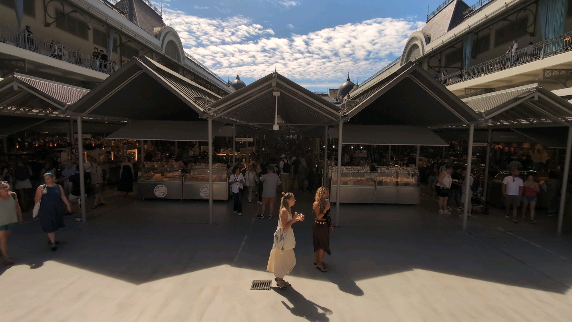 This is a street-level, wide-angle shot of the Mercado do Bolhão in Porto, Portugal, on a sunny day with a partially cloudy sky. The market has undergone renovation and now features a modern, open-air section with a concrete floor and peaked, dark-colored awnings supported by metal poles. The market stalls are visible in the background, showcasing various goods.
In the foreground, a woman in a light-colored dress and a woman in a brown skirt and dark top are walking and talking. To the left, another woman in a dark blue dress is walking with a white bag. Further left, a woman in a green top and shorts is also walking. Several other individuals are seen walking in the middle ground and background, some browsing at stalls and others conversing.
The architecture of the building frames the scene, with upper levels featuring balconies and arched windows, suggesting a historic structure that has been updated. The sunlight casts strong shadows on the concrete ground, indicating a bright day. The overall atmosphere appears to be one of a bustling public market.