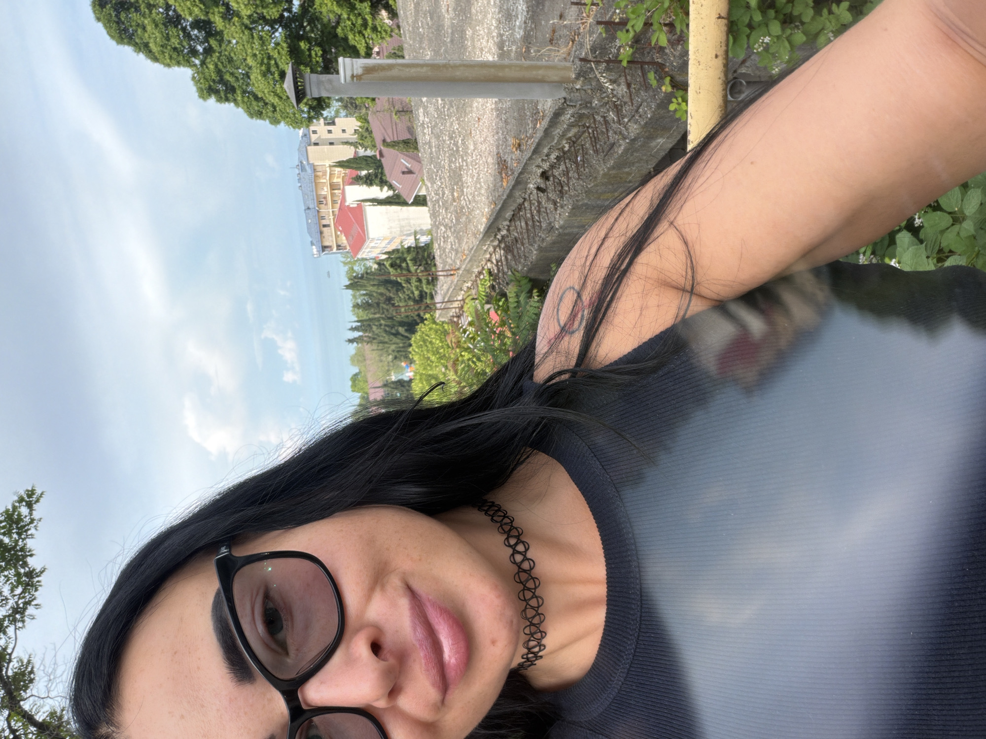 A woman with dark hair and glasses is taking a selfie outdoors in Sochi, Russia. She is wearing a black tank top and a choker necklace. In the background, there are buildings, trees, and the sea. The sky is cloudy.