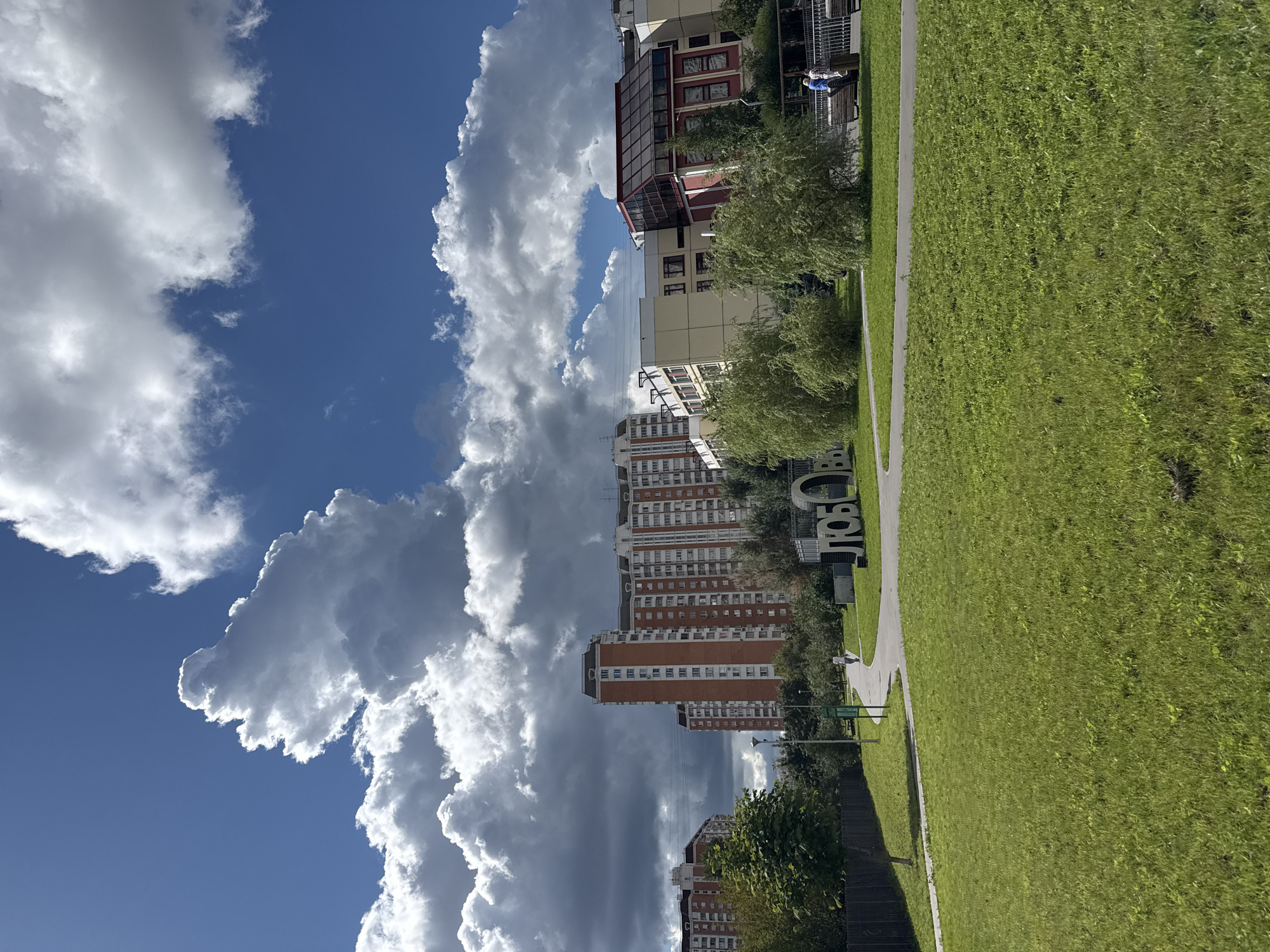 The image depicts a sunny day in a park in Lyubertsy, Russia, with dramatic clouds in a bright blue sky. In the background, several tall residential buildings with orange and white facades are visible, alongside a modern building with a red and glass facade. A large white sculpture spelling out "ЛЮБО" (Lyubo, meaning "love" in Russian) stands in the middle ground. A paved pathway curves through a lush green lawn dotted with trees. Two individuals are seated on a bench near the sculpture, and another person walks along the path. The overall scene suggests a pleasant urban park environment.