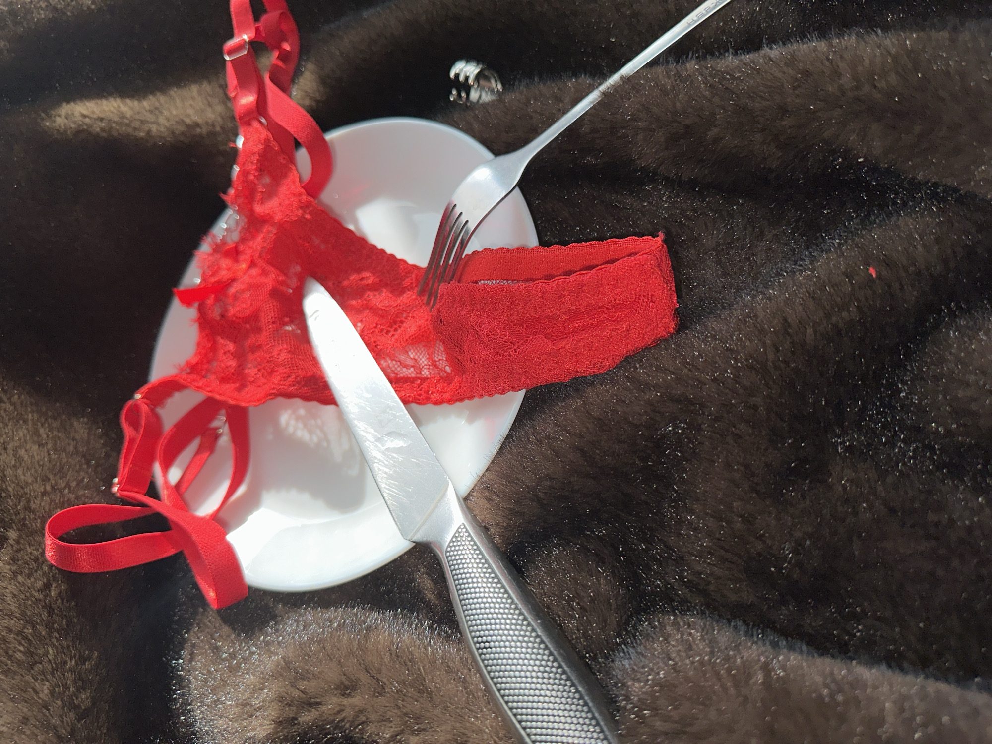 A white plate with a red lace thong, a fork, and a knife is positioned on a dark brown, plush surface that resembles fur or velvet. A ring is visible on the plate to the side of the thong. The photograph appears to be taken indoors. The location in Chaikovsky, Russia is not discernible from the image.