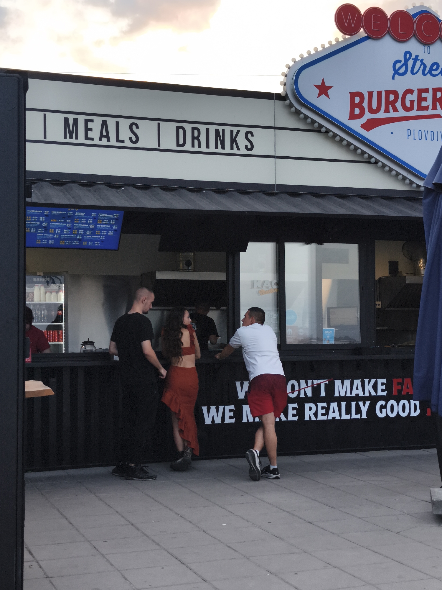 The image captures an outdoor scene in Plovdiv, Bulgaria, featuring a burger joint named "Street Burger." The establishment has a prominent sign with the word "WELCOME" in red circles at the top, followed by "TO Street BURGER" in stylized blue and red lettering. Below the sign, there is a black facade with white lettering that reads "MEALS | DRINKS." Further down, a black banner displays the text, partially obscured, "WE DON'T MAKE FA..." and "...WE MAKE REALLY GOOD...".
In the foreground, three individuals are visible. On the left, a man in a black t-shirt and dark pants stands facing away from the camera, looking towards the burger joint. In the center, a woman with long dark hair wears a red crop top and a flowing orange skirt, facing the man and the establishment. To the right, another man in a white t-shirt and red shorts is seen from the side, leaning slightly forward and looking towards the counter.
Behind the counter, through the windows of the burger joint, glimpses of the interior can be seen, including refrigerators and kitchen equipment. A blue umbrella or shade is partially visible on the far right of the image. The ground is paved with square tiles. The sky above is partly cloudy with hints of sunset colors. A menu board is visible inside the establishment, displaying various burger options and prices.