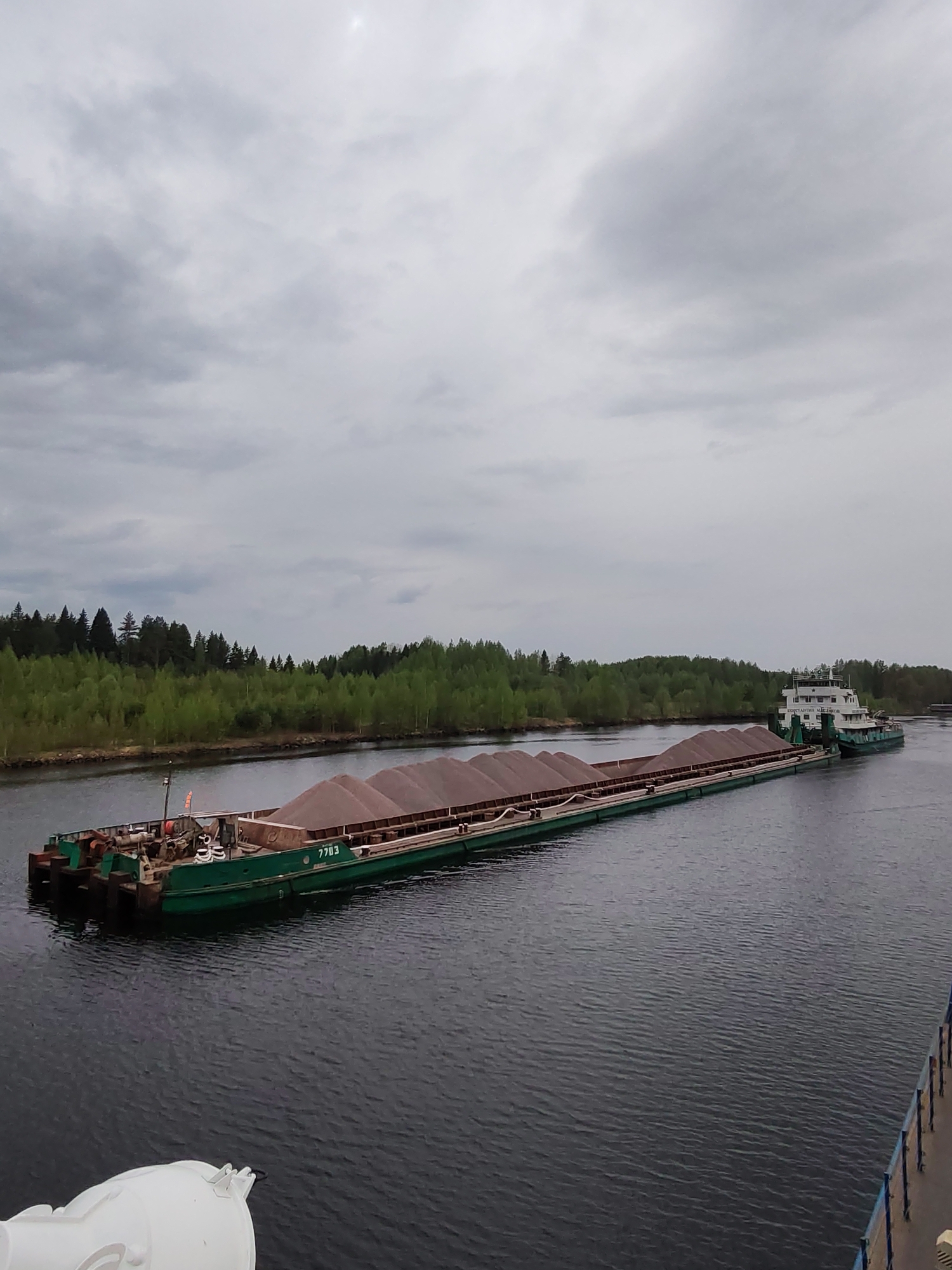 The image shows a barge being towed on a waterway, likely a canal, in an area with overcast skies. A long, green barge filled with what appears to be gravel or sand is being towed by a white and green tugboat. The barge is marked with the number "7703". The scene is set on a body of water with rippled surface. Trees line the edge of the waterway on one side. The photo is taken from a slightly elevated perspective, potentially from another vessel or structure near the water. It is most likely taken in the vicinity of Alekseevskoe, Russia.