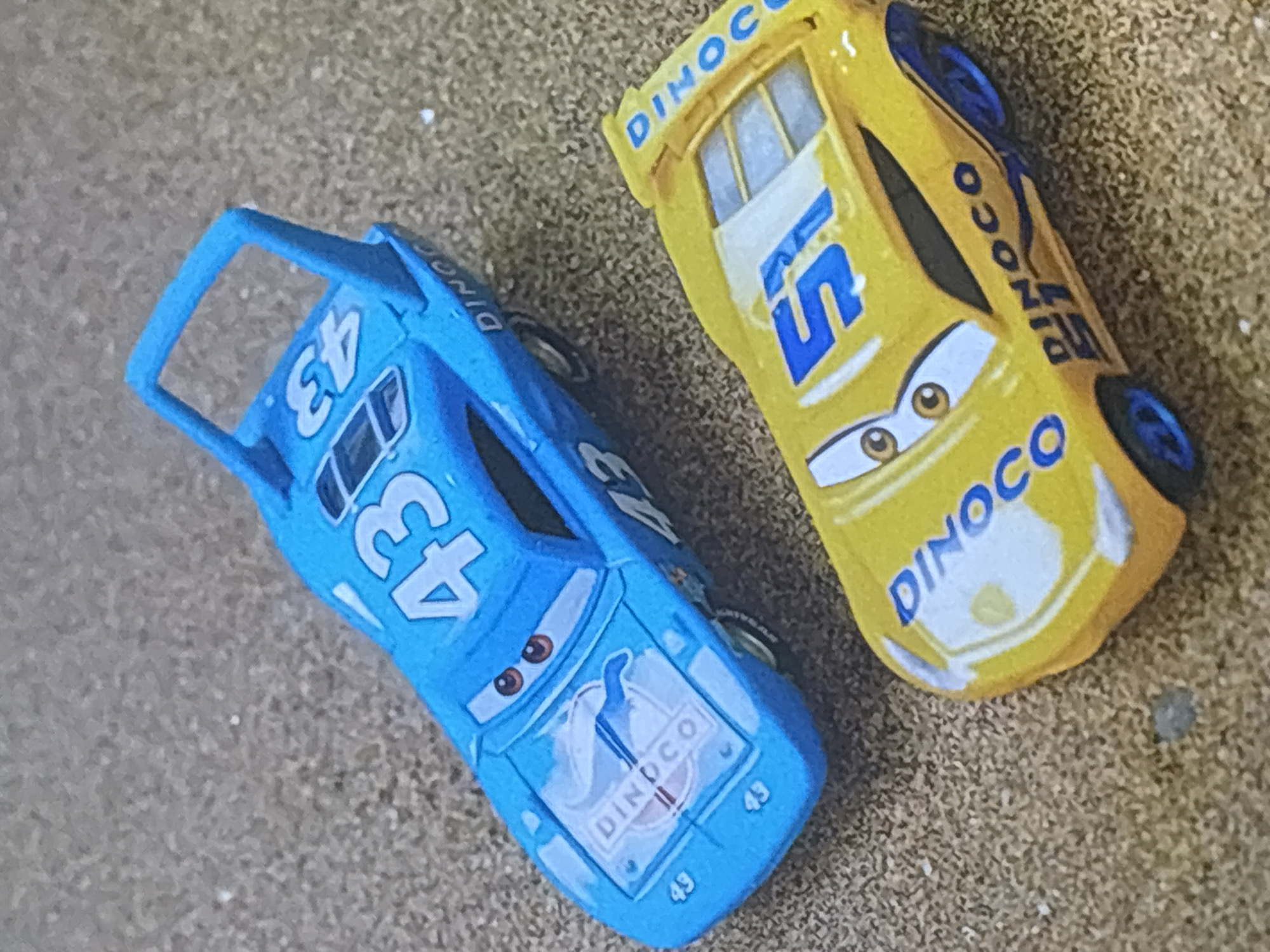 Two toy cars, a blue one with the number 43 and a yellow one with the number 51, are displayed on a sandy surface. The blue car is a replica of "The King" from the movie Cars, and the yellow car is a replica of Lightning McQueen. Both cars are depicted with their headlights on and their racing decals visible. The sandy surface is textured, suggesting an outdoor environment. The image is cropped from a larger scene and does not contain any information about the city of Dubai or the United Arab Emirates.