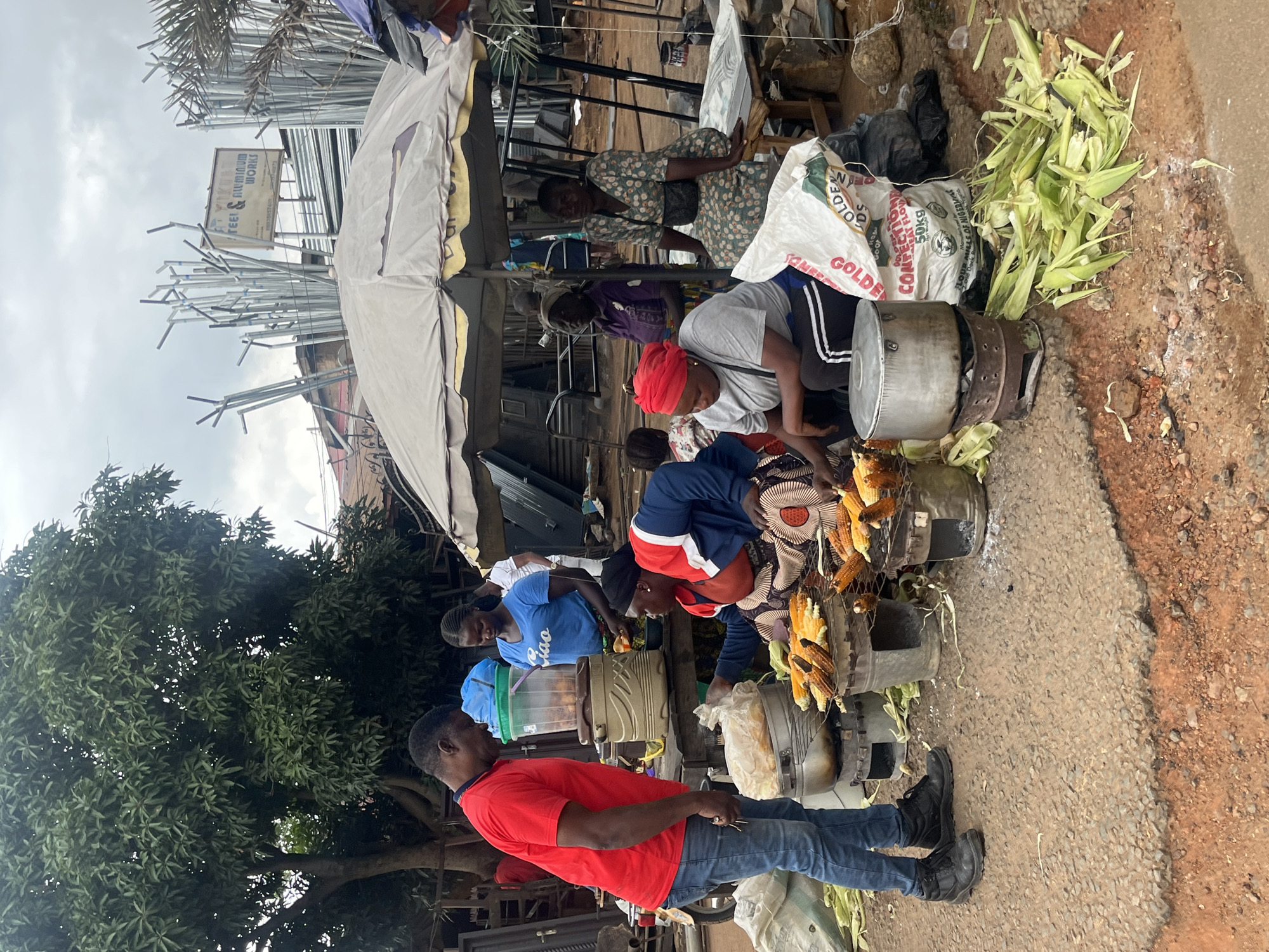 The image depicts an outdoor market scene in Gura Top, Nigeria. Several individuals are present, some engaged in selling food, others as customers. A prominent stall features grilled corn on the cob displayed on metal racks over a cooking apparatus. Nearby, sacks of what appear to be grain or flour are visible, along with discarded corn husks.
The people in the scene are dressed in casual clothing. One woman wears a red headscarf and a grey shirt, interacting with the corn stall. Another woman in a blue t-shirt stands beside a container of food. A man in a red t-shirt and blue jeans is also present, looking towards the market.
The setting is a bustling street market, with a mix of makeshift stalls and temporary structures. Scaffolding or metal pipes are visible in the background, suggesting ongoing construction or market setup. The ground is a mixture of dirt and asphalt, with some areas appearing wet. A large leafy tree provides some shade.
The time of day appears to be daytime, possibly morning or afternoon, judging by the lighting. The sky is overcast, indicating potentially cloudy weather. The overall atmosphere seems busy and active, typical of a market environment.
Visible text includes "GOLDEN" on a white sack and "50KG" on another sack. A sign in the background is partially visible with "STEEL & ALUMINUM WORKS" and a phone number.