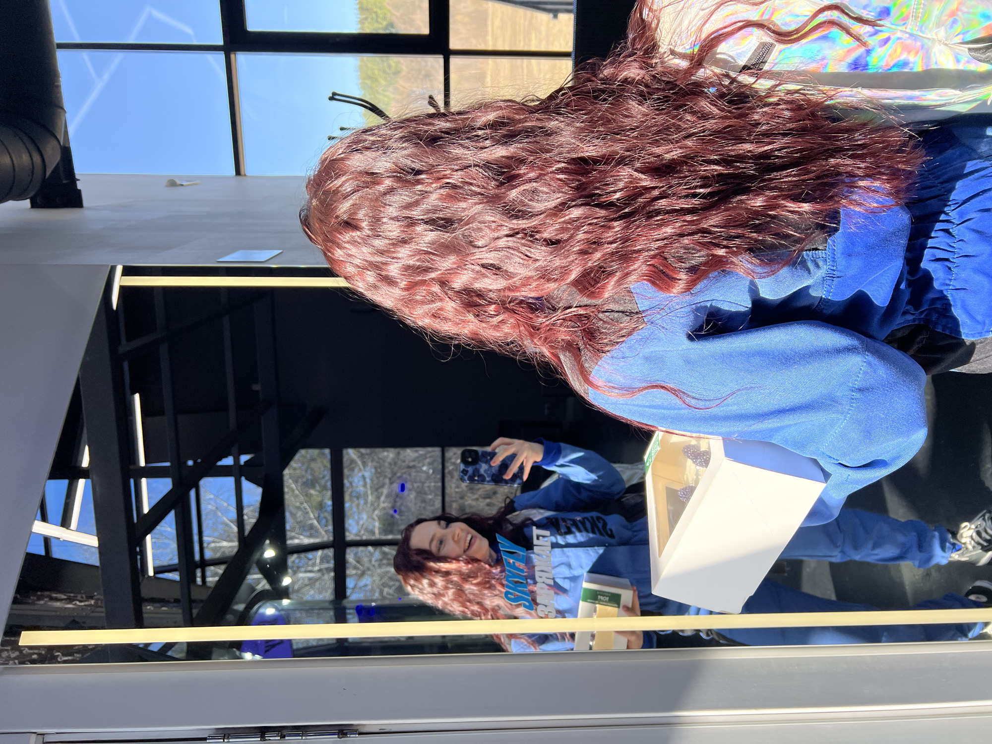 A young woman with long, wavy, auburn hair is standing indoors, taking a selfie in a large mirror. She is wearing a bright blue tracksuit. She is holding a white rectangular box in her left hand and a cell phone in her right hand, presumably to take the photograph. The mirror reflects her image and the scene behind her, showing large windows with a view of trees. The location is not discernable in the image.