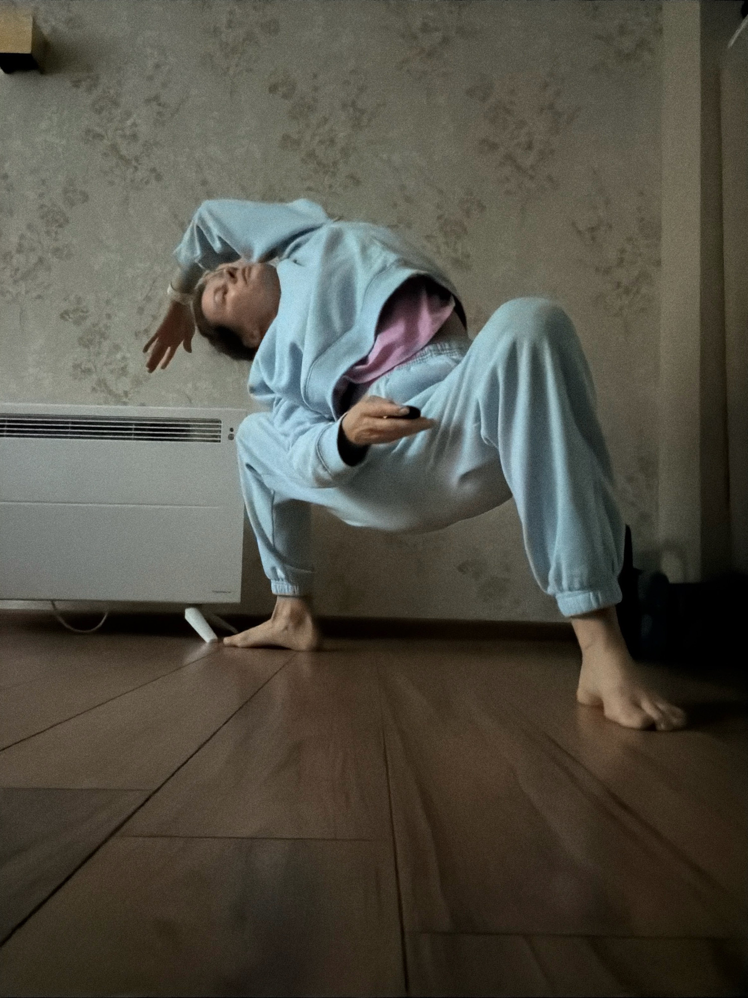 An individual is performing a backbend on a wooden floor in front of a white electric heater. The individual is wearing a light blue hooded sweatshirt and matching sweatpants. Their body is arched backward, with their head tilted back and their left arm extended, palm facing up. They are holding a dark object, possibly a remote control, in their right hand. The wall behind them is covered in a patterned wallpaper. The scene appears to be indoors, in a residential setting, potentially an apartment in Riga, Latvia.