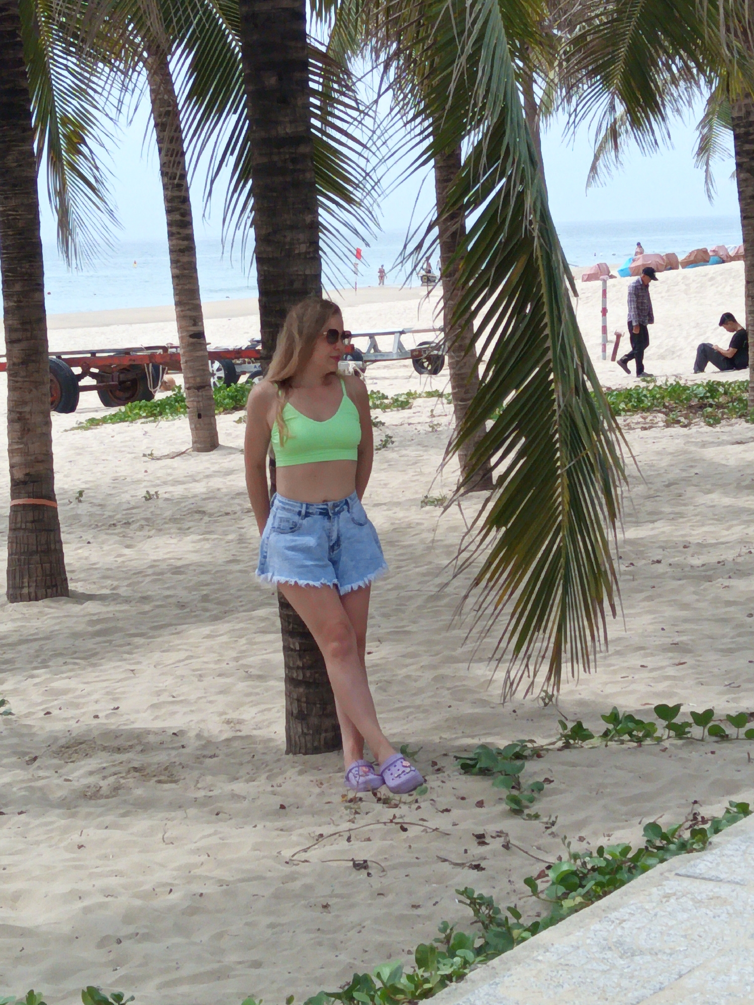 A young woman with blonde hair stands on a sandy beach in Da Nang, Vietnam, leaning against a palm tree. She is wearing a neon green crop top and light blue denim shorts, with purple sandals on her feet. The beach is lined with palm trees, and in the background, the ocean is visible with a few people in the water and on the shore. There is also a red trailer with large wheels on the sand. Other people are visible in the distance, including a man in a plaid shirt and a man sitting on the sand. The weather appears to be sunny.