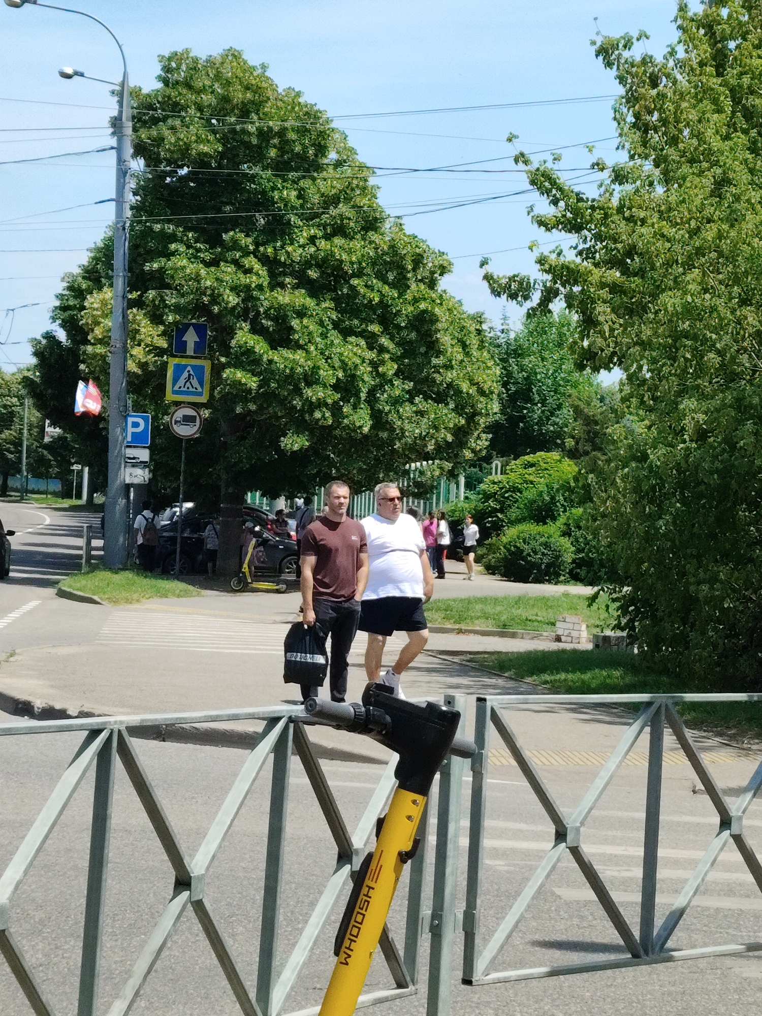 The image captures a sunny day scene in Krasnodar, Russia, likely on a street with moderate pedestrian and vehicle traffic. In the foreground, a metal railing with a diamond pattern is visible, with a yellow electric scooter parked beside it. The scooter has "WHOOSH" written vertically on its stem. Two men stand on a crosswalk in the middle ground, facing towards the viewer and slightly to the right. The man on the left, wearing a maroon t-shirt and black pants, holds a black bag. The man on the right, in a white t-shirt and black shorts, is looking away from the camera. Behind them, several cars are parked, and other pedestrians are visible further in the background, along with trees and utility poles. Traffic signs, including a pedestrian crossing sign and a "no trucks" sign, are also present. The overall environment suggests a typical urban setting in Krasnodar during warmer weather.