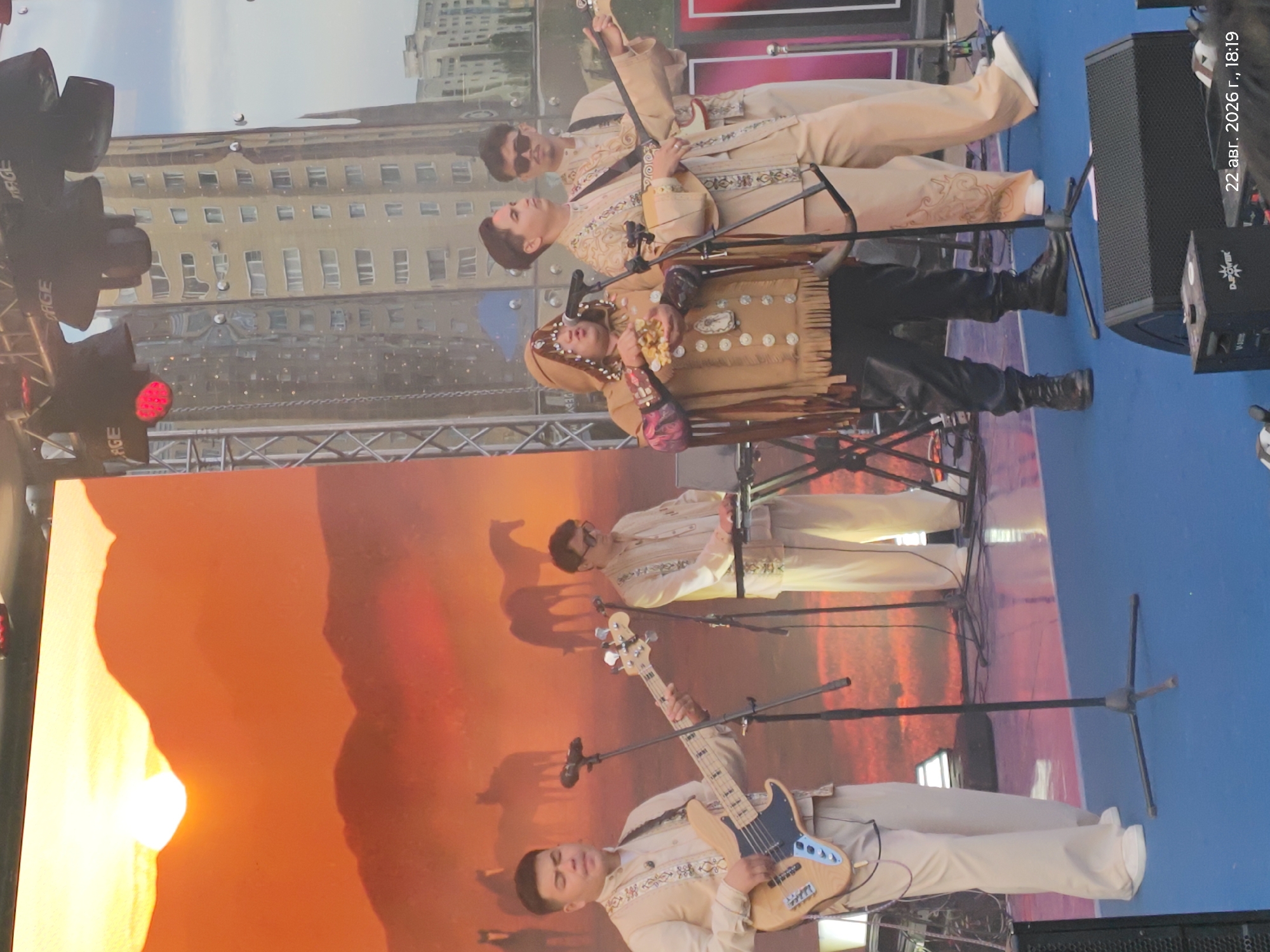 On an outdoor stage with a large screen displaying a desert landscape with horses and a setting sun, a band performs. The screen's backdrop is a warm orange, depicting silhouetted horses against a hazy sky. In the foreground, four individuals are visible. On the left, a man in a cream-colored outfit with embroidered details plays a bass guitar. To his right, another man in a similar cream outfit stands behind a keyboard. Center stage, a performer in a distinctive brown, hooded outfit with fringe and numerous circular adornments holds something in their hands and sings into a microphone. To the right of the singer, a man in a cream outfit plays a stringed instrument, possibly a dombra, and wears sunglasses. The stage floor is a vibrant blue, and black speakers are positioned at the front. In the background, a reflection on a surface shows tall, light-colored buildings, characteristic of the city of Astana, Kazakhstan. A timestamp in the lower right corner indicates "22 авг. 2026 г., 18:19," suggesting the photograph was taken on August 22, 2026, at 6:19 PM.