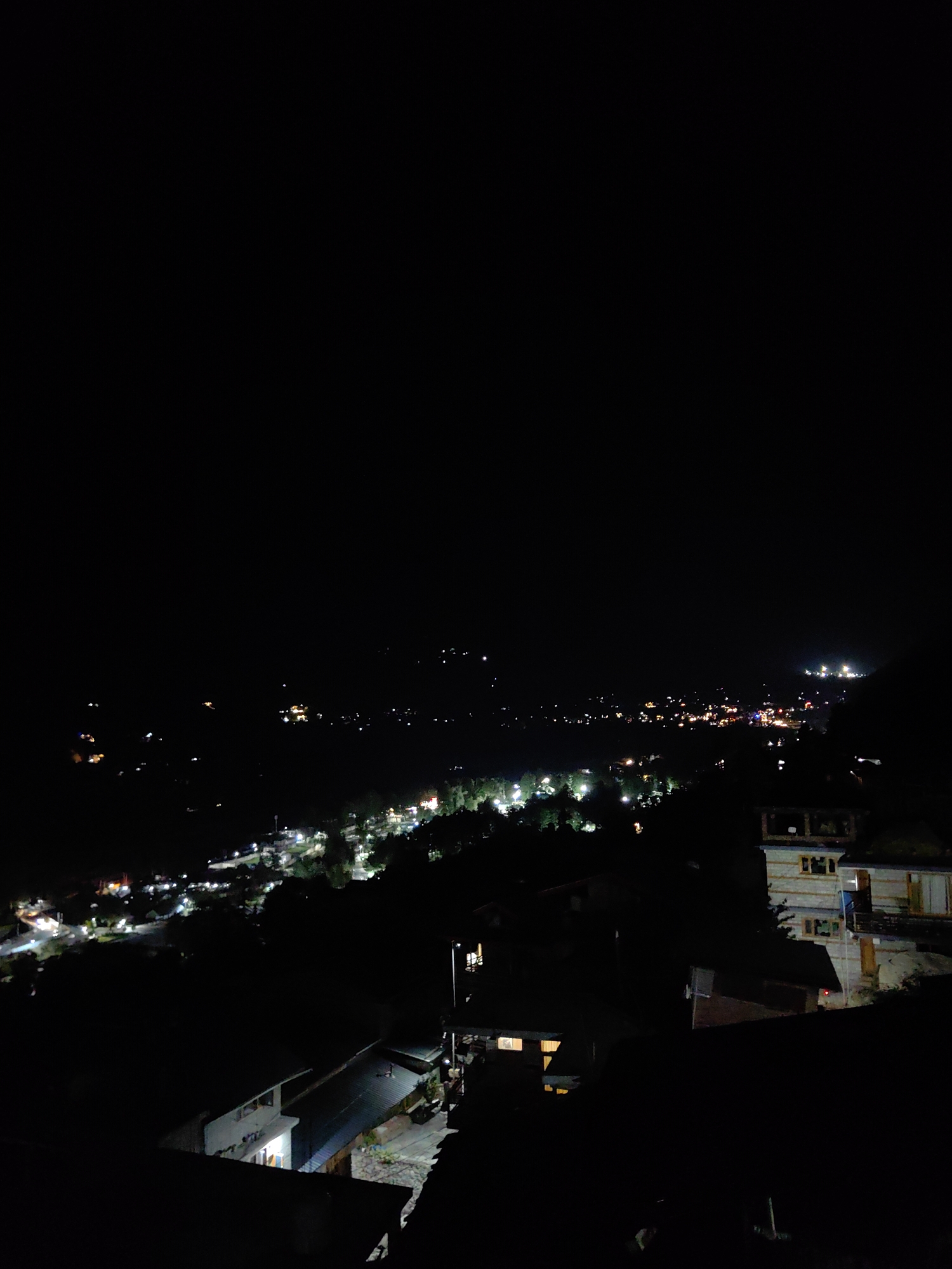 The image depicts a nighttime aerial view of a town, likely Vashisht, India, nestled within a hilly terrain. The landscape is dominated by darkness, punctuated by the warm glow of numerous lights emanating from buildings and streetlamps. In the foreground, several structures with lit windows are visible, suggesting residential or commercial buildings. One building in the mid-ground on the right appears to be a multi-story structure with balconies and windows, also illuminated. The middle ground reveals a denser cluster of lights, indicating the heart of the town, with roads and pathways likely visible through the illumination. Further in the distance, more lights stretch across the landscape, hinting at the town's extent. The presence of trees is subtly suggested by darker masses interspersed with the lights. The overall scene conveys a sense of a tranquil, illuminated settlement under the cover of night, characteristic of a mountainous town.