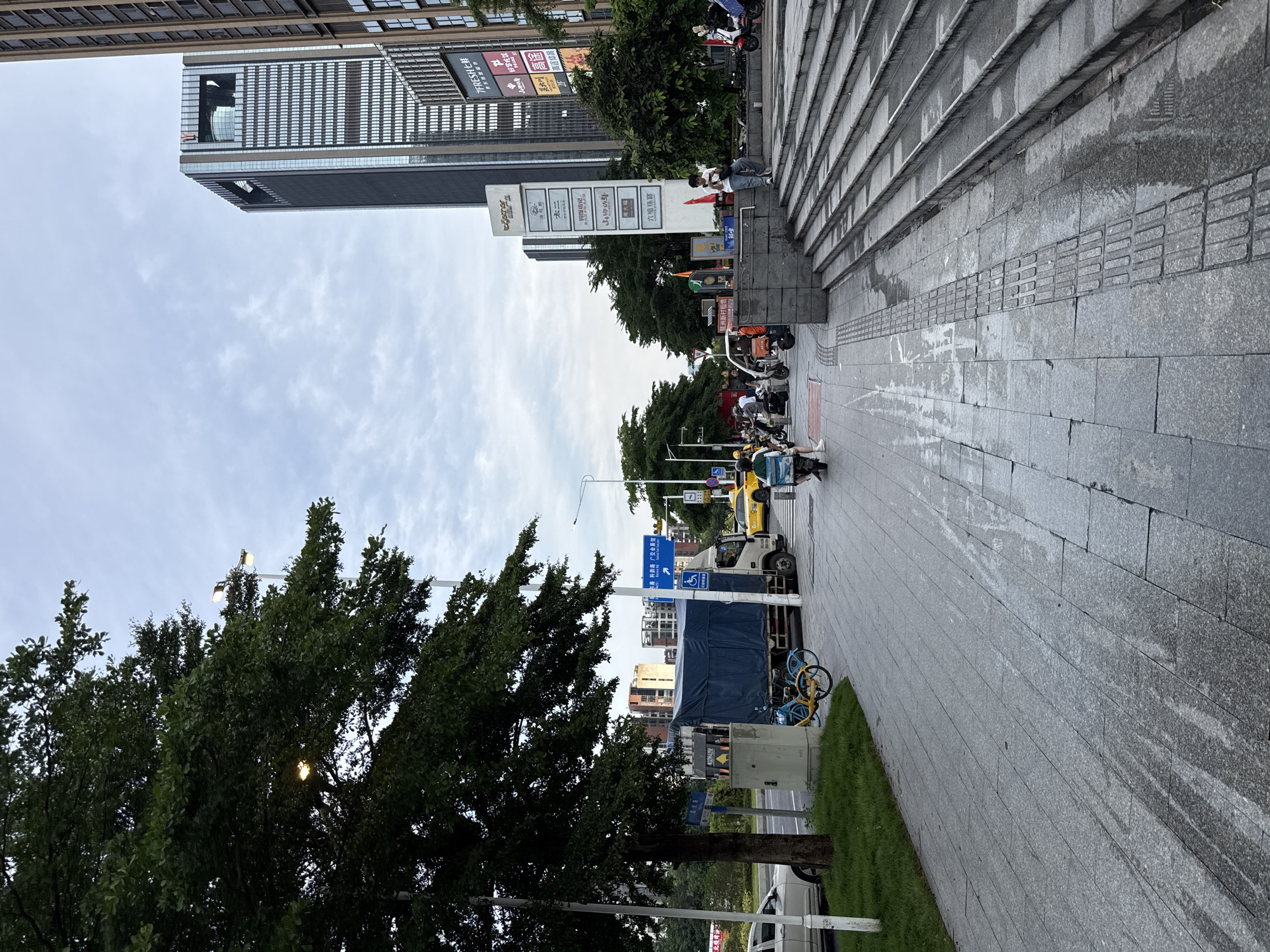 This is a vertical, outdoor photograph taken in Haizhu District, China. The scene depicts a street with a sidewalk and a large set of stairs. Several people are visible, some sitting on the stairs and others on scooters or bicycles. There's a variety of vehicles, including scooters, bicycles, a yellow taxi, and a truck covered with a blue tarp.
The background features modern architecture, including a tall, sleek skyscraper. A sign with multiple panels displays various Chinese characters and logos, including "7FRESH七鲜", "太二", "高途", and "六福珠寶". The lighting suggests it is daytime, possibly late afternoon or early evening, with overcast or cloudy skies. The pavement appears damp, indicating recent rain or cleaning. The presence of green trees and grass provides a touch of nature amidst the urban environment. The overall atmosphere seems relatively calm, with individuals engaged in their daily activities.