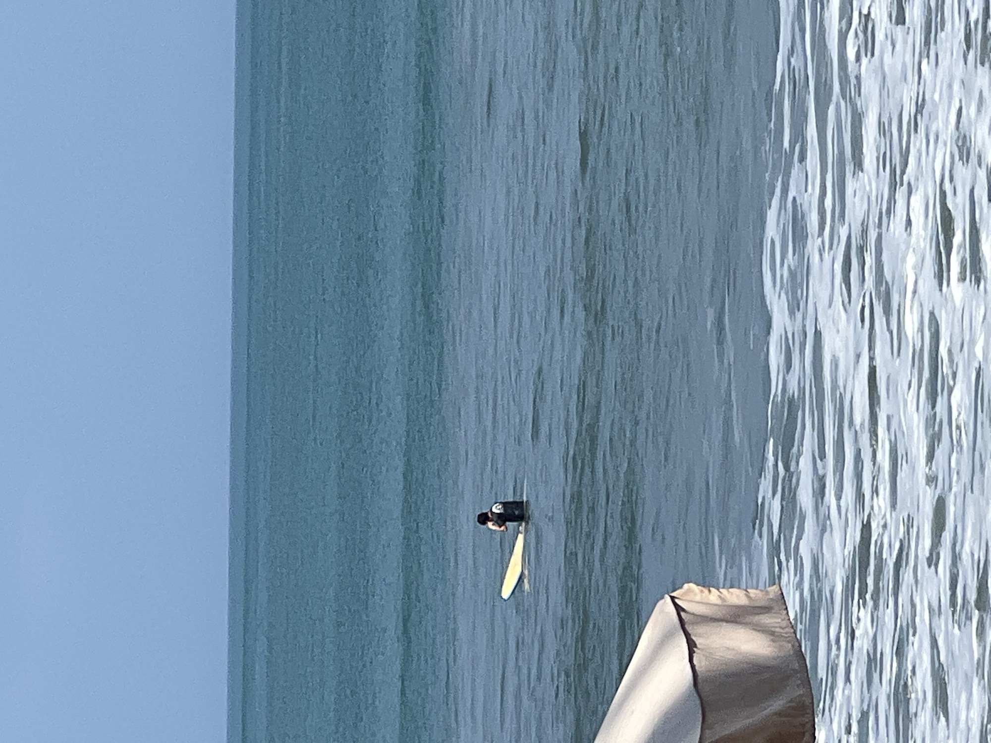 The image depicts a serene ocean scene with a person standing in the water next to a yellow surfboard. The person is wearing a dark-colored rash guard. The water is a calm, deep blue, with small waves breaking in the foreground, creating white foam. In the lower left corner, a portion of a light-colored beach umbrella is visible, casting a shadow on the water. The horizon is clearly defined, separating the deep blue sea from a pale blue sky. The overall atmosphere is tranquil and suggests a relaxed day at the beach. This scene is consistent with the coastline of Kuta, Indonesia, known for its beaches and surfing.