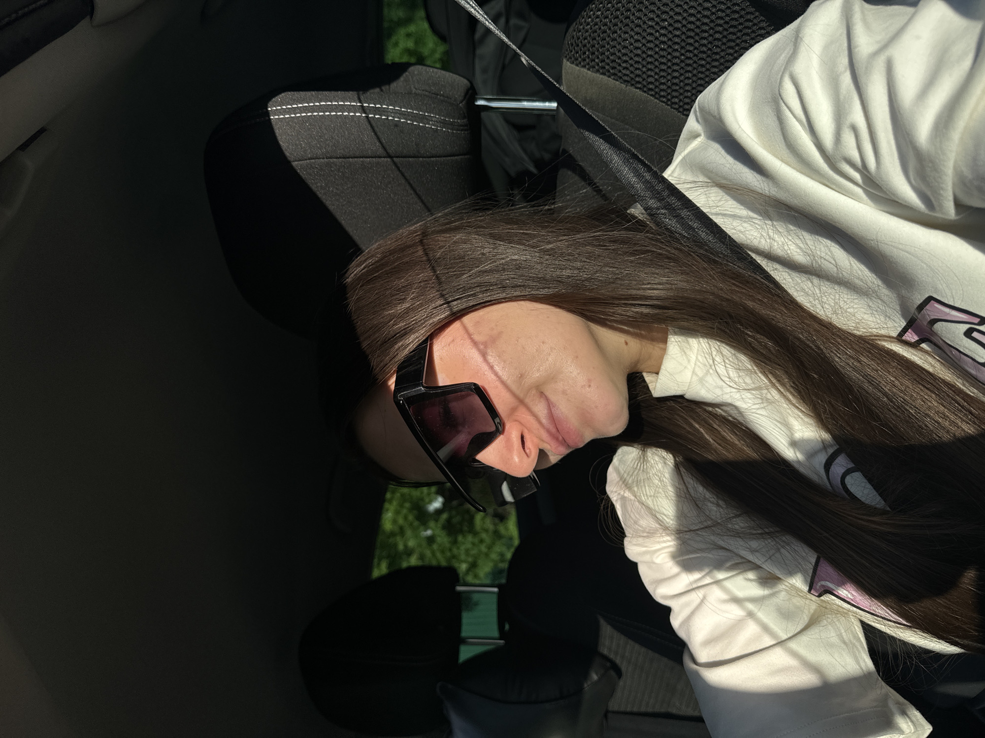 The image shows a person sitting in the passenger seat of a car. The person has long, dark brown hair and is wearing sunglasses and a white long-sleeved shirt. A seatbelt is fastened across their body. The interior of the car is visible, including the black seat and the dashboard in the background. Outside the car window, green vegetation can be seen. The image does not provide enough information to determine the location, or any information about Sharapovka, Russia.