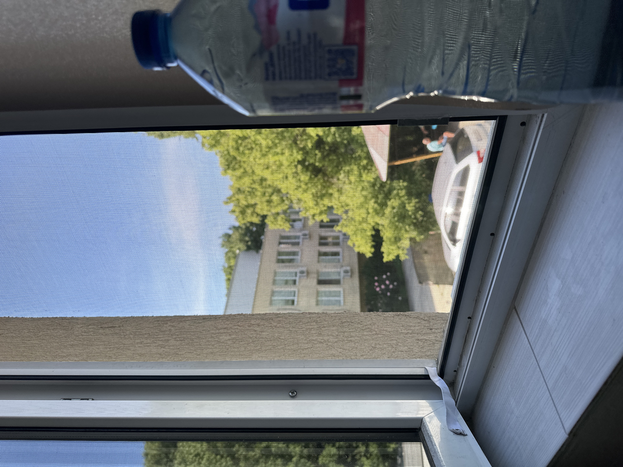 The view from an open window with a screen shows a building with multiple windows, green trees, and a parked white car. A water bottle rests on the window frame. A second window is visible below the first, showing more trees. This scene is likely in a residential area of Sofia, Bulgaria, given the architecture and vegetation typical of the city.