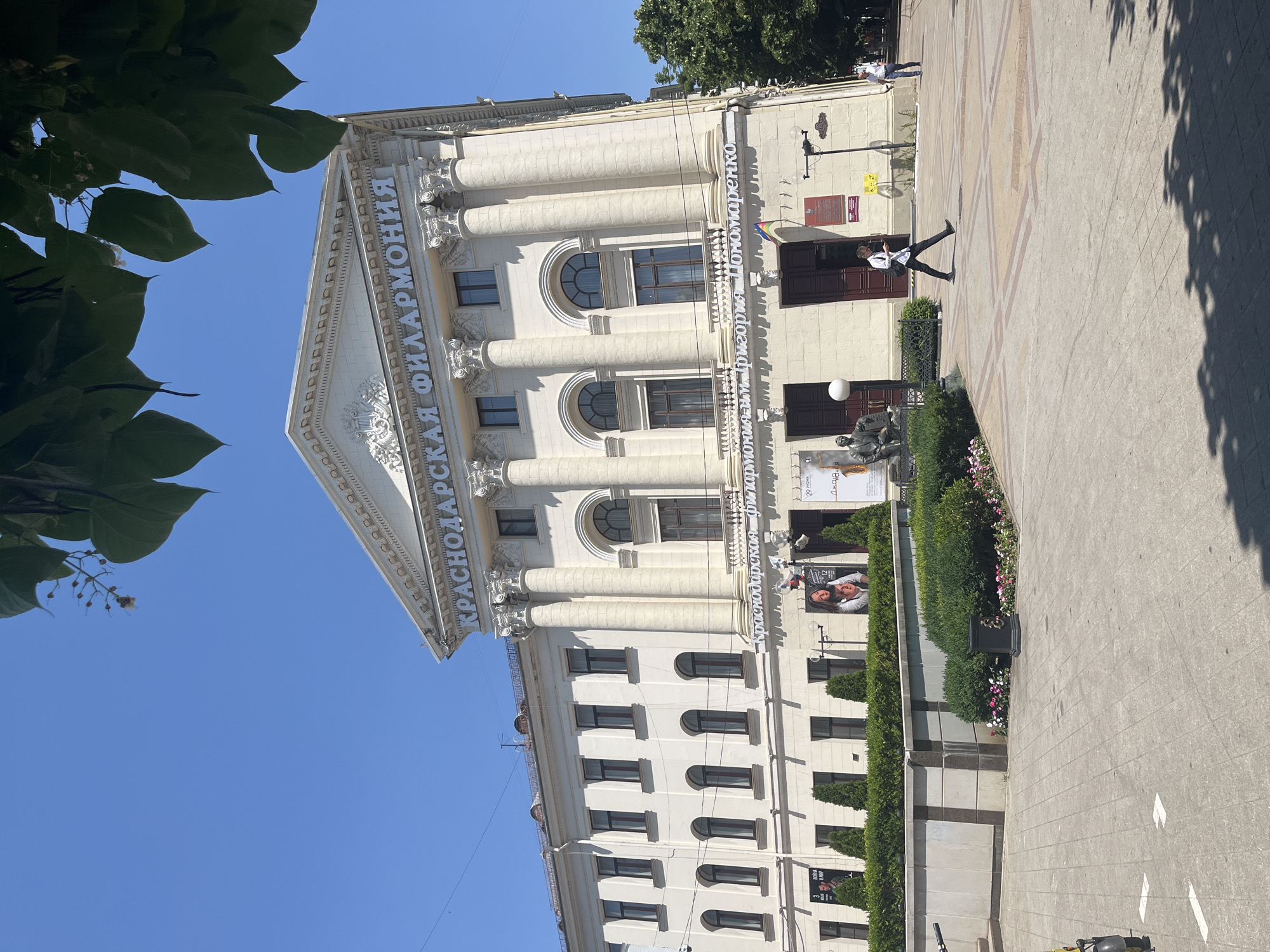 The image depicts the Krasnodar Philharmonic Hall, a grand building with classical architecture. The facade is adorned with columns and arched windows. In front of the building, a paved area is visible, with a person walking across it. The time of day appears to be daytime, with a clear blue sky and bright sunlight casting shadows, suggesting fair weather. Green foliage from trees and bushes surrounds the building and the paved area.
Visible text on the building reads "КРАСНОДАРСКАЯ ФИЛАРМОНИЯ" (Krasnodar Philharmonic) and "имени Григория Пономаренко" (named after Grigory Ponomarenko). There are also posters on the building, one of which shows two women. The overall scene conveys a sense of cultural and urban activity on a sunny day.
