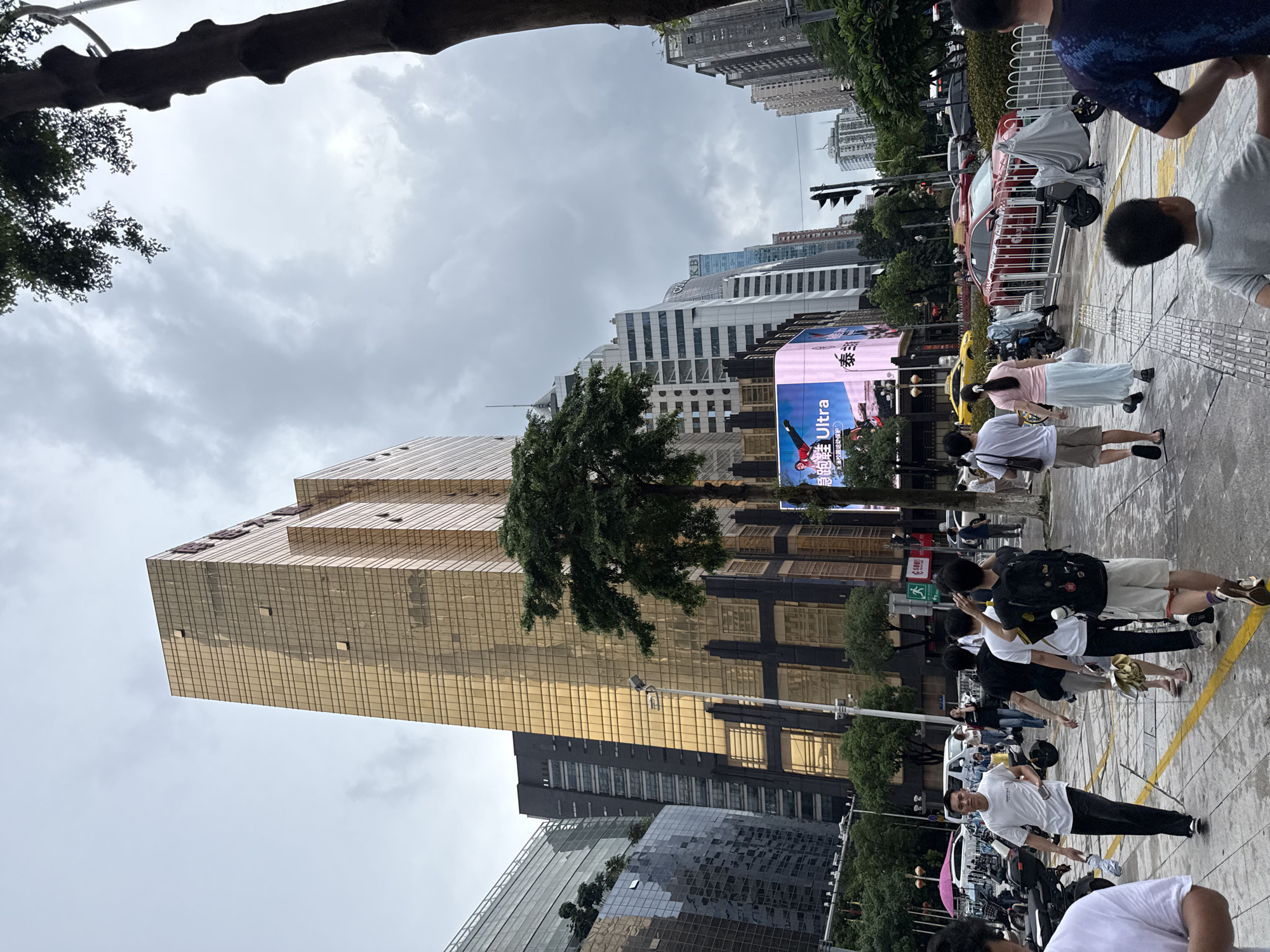 In Tianhe District, China, a cloudy day presents a street scene dominated by a striking golden skyscraper. The building's facade is a grid of reflective glass panels, reflecting the overcast sky. To its left, a darker, modern building with numerous windows rises, also a prominent feature of the cityscape. In the foreground, a sidewalk is populated by several individuals walking in various directions. A man in a white t-shirt and black pants walks towards the viewer, holding a phone and a bottle of water. Behind him, a group of men in casual attire walks away. Some are carrying backpacks, and one is holding a yellow umbrella. Across the street, traffic is visible, including a red car and several motorcycles parked along the sidewalk, some covered with tarps. A large digital billboard displaying an advertisement for "Ultra" shoes is mounted on a building to the left of the golden skyscraper. The overall environment suggests an urban setting with commercial activity and pedestrian traffic.