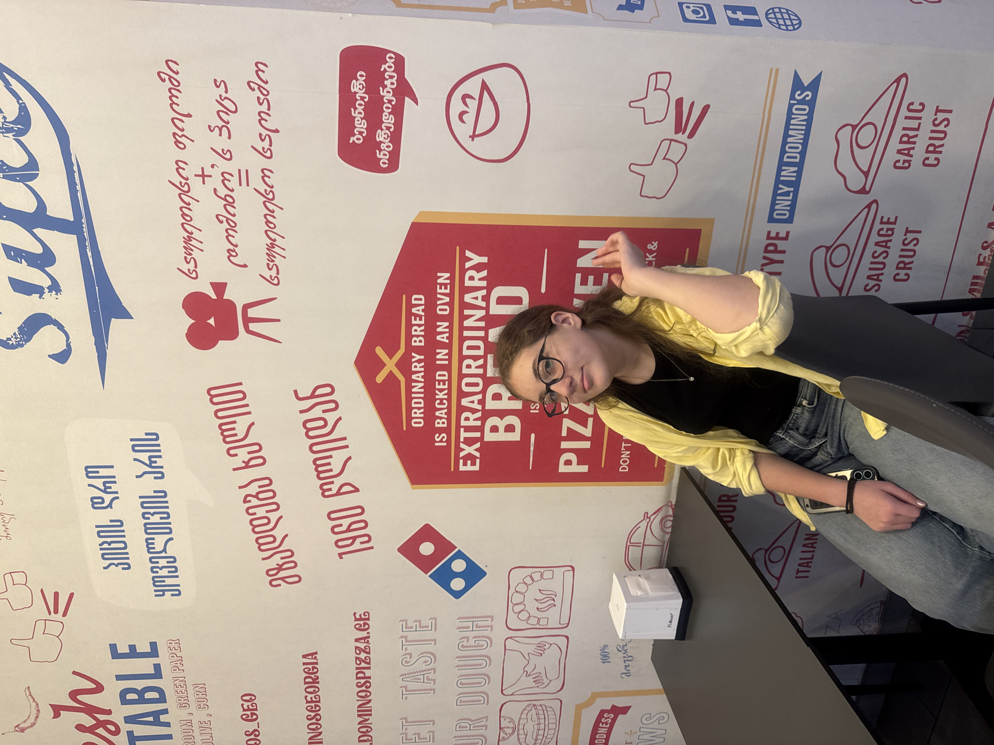 This is a medium shot, eye-level, indoor photograph taken at a Domino's Pizza restaurant in Tbilisi, Georgia. The background is a wall with graphics and text related to Domino's Pizza. In the foreground, a young woman with light brown hair and glasses is seated at a black table, facing the camera. She is wearing a yellow cardigan over a black top and blue jeans. Her right arm is resting on the back of the chair, and her left hand is partially visible. The wall behind her features various slogans and illustrations, including the Domino's logo, images of pizza, and Georgian text. A small white napkin dispenser is on the table to her left. The overall lighting is moderate, and the focus is on the woman and the restaurant's branding.