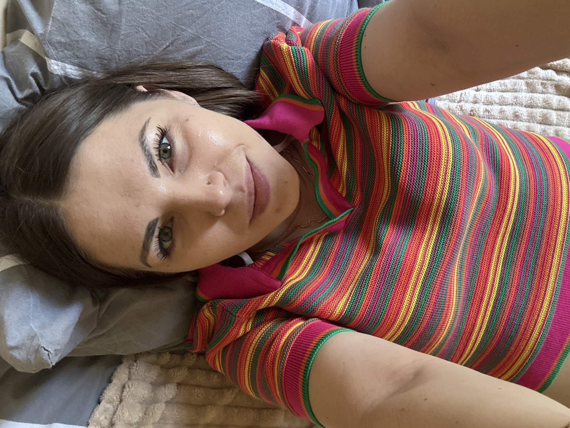 A woman with light skin, brown hair, and green eyes lies on a gray pillow on a beige textured blanket. She is wearing a striped top with pink, red, orange, yellow, and green colors and a pink collar. Her gaze is directed towards the camera, with a slight smile on her face. A nose ring is visible on her left nostril. The location in the city of Раздолье, Russia is not determinable from the image.