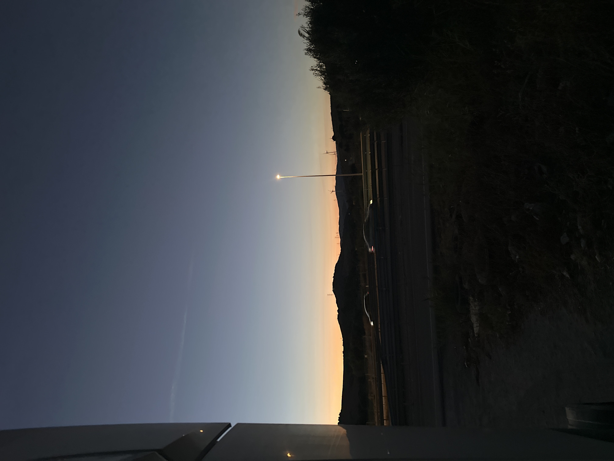 The image depicts a twilight scene on the outskirts of Los Barrios, Spain, likely near a highway. The sky transitions from a deep blue at the top to a warm orange glow near the horizon, indicating either sunrise or sunset. Silhouetted against this vibrant sky are rolling hills, topped with what appear to be wind turbines and possibly some communication towers.
In the foreground, a highway is visible, with two cars moving in the same direction, their headlights illuminated. The road is bordered by a concrete barrier and some low-lying vegetation. To the far left, a portion of a white vehicle, possibly a camper van or RV, is visible, suggesting the vantage point might be from within or beside such a vehicle. The ground in the immediate foreground is dark and appears to be unpaved, with rough terrain and scattered debris. To the right of the highway, a dense patch of dark foliage forms a shadowed area, obscuring details within it. A single streetlamp stands illuminated along the highway, casting a cone of light. The overall atmosphere is serene and transitional, capturing the quiet moments between day and night.