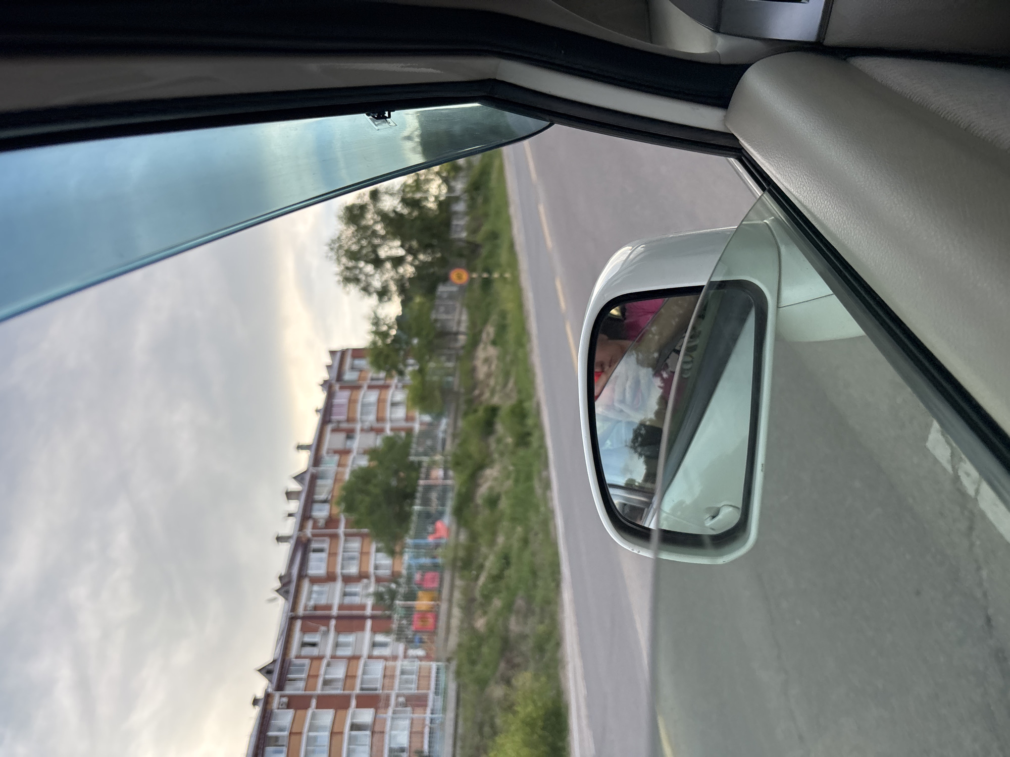 An exterior shot captures a side view from within a car, looking out the passenger window. A white side-view mirror reflects a person wearing a pink top and a portion of what appears to be the car's interior. Beyond the mirror, a paved road stretches into the distance, bordered by a grassy embankment. In the background, a large, multi-story apartment building with a reddish-orange facade and white-framed windows stands prominently. Trees dot the landscape between the road and the building. The sky is overcast with visible clouds. The scene is set in Plodopitomnik, Russia.